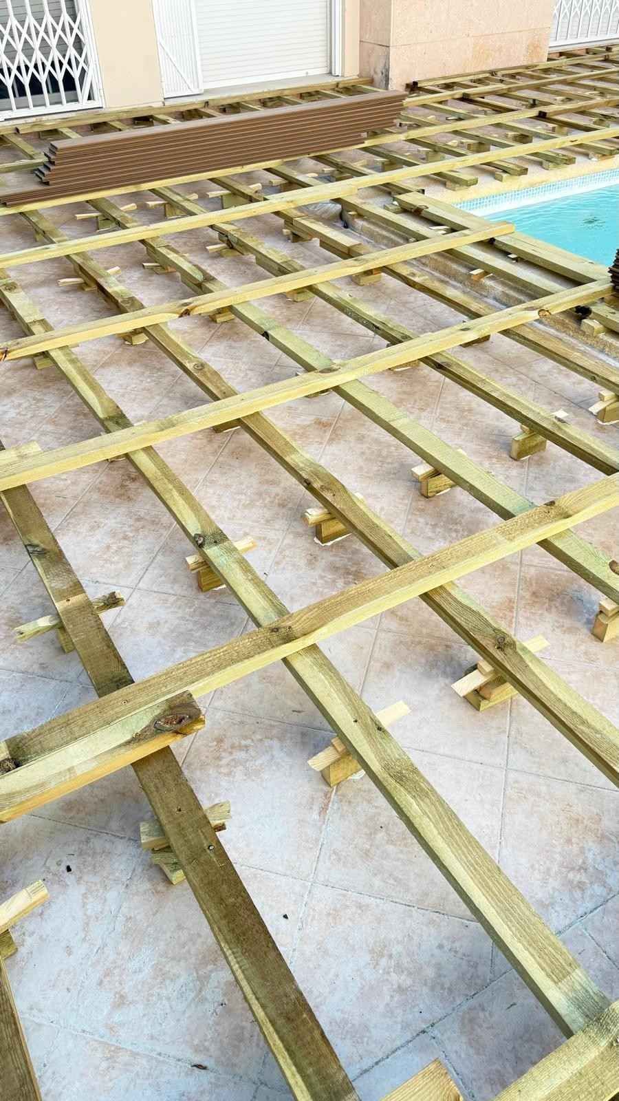 Estructura de madera para terraza en construcción junto a una piscina, con vigas amarillas sobre hormigón.