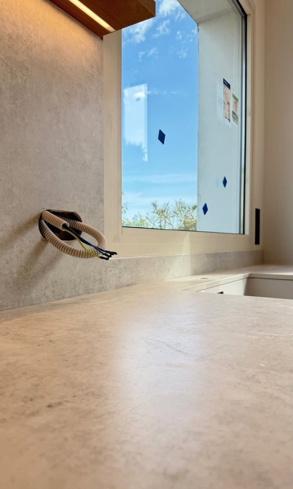 Encimera de piedra junto a un lavabo y un espejo de baño, con un cielo azul reflejado en la ventana.