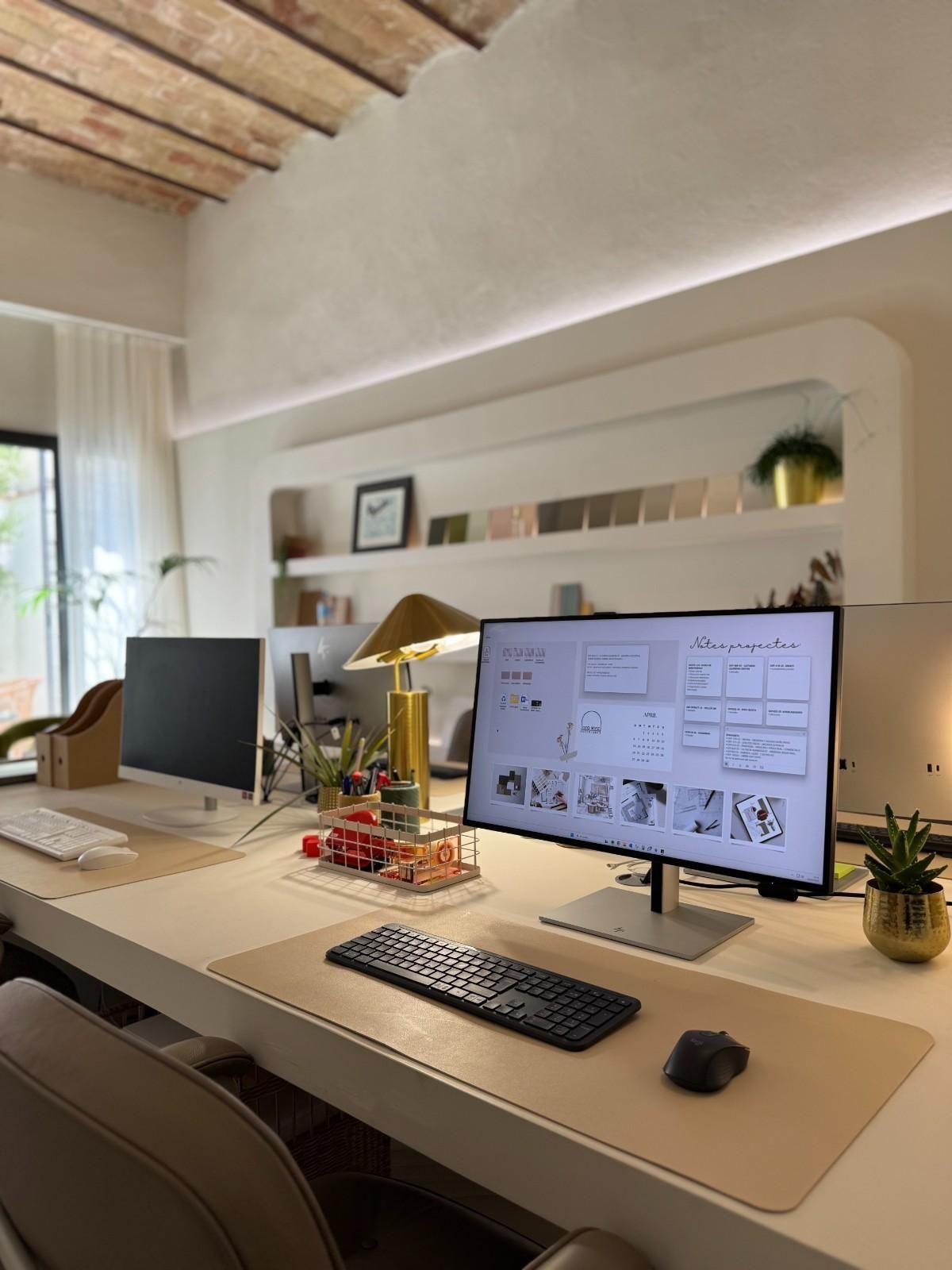 Escritorio moderno para oficina en casa con dos monitores, teclado, lámpara, libros y plantas bajo un techo abovedado.