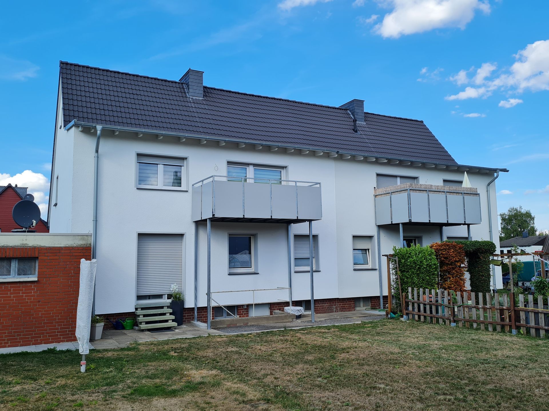 Weißes zweistöckiges Haus mit grauem Dach, Balkonen und einem umzäunten Garten unter blauem Himmel