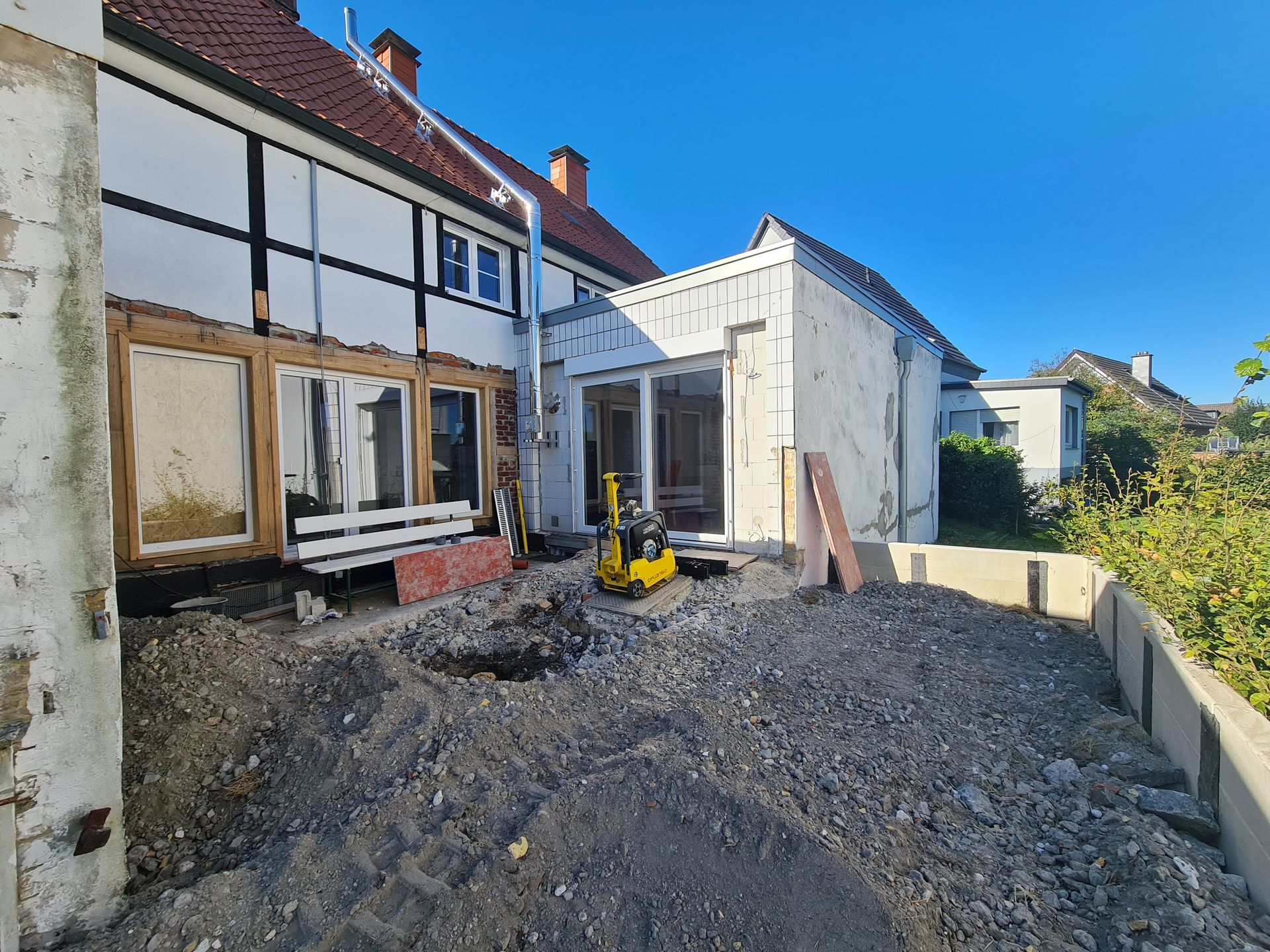 Eine Baustelle im Hinterhof mit ausgehobener Erde, einem gelben Bagger und einer Hausfassade, die renoviert wird.