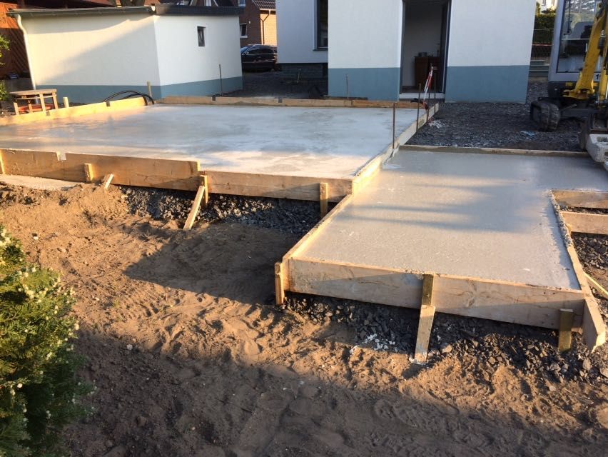 Eine im Bau befindliche Beton-Terrasse neben einem Haus, deren Ränder unfertig sind und die von Erde umgeben ist.