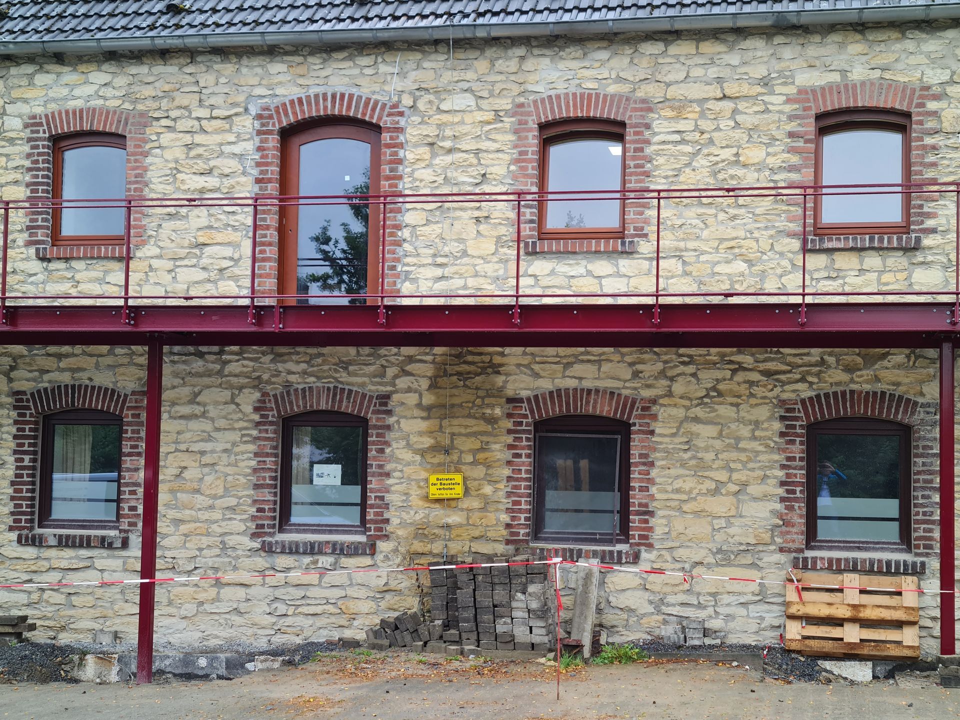 Alte Steinfassade mit rot umrandeten Fenstern und Türen, rotem Metallgeländer und einem Stapel Holzpaletten.