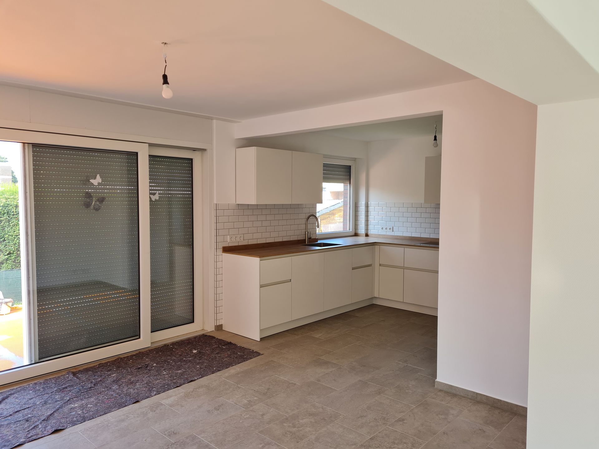 Leere, moderne Küche mit Essbereich, weißen Schränken, Holzarbeitsplatten, Glasschiebetüren und grauem Boden.
