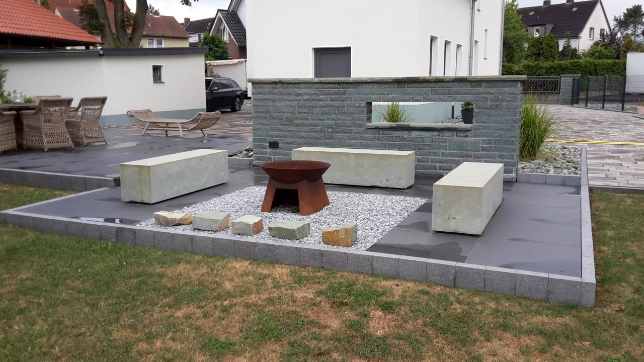 Moderner Sitzbereich im Freien mit grauen Bänken, Feuerstelle und gepflasterter Terrasse in einem Wohngarten