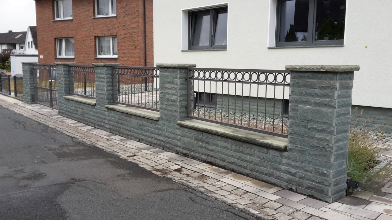 Stein- und Metallzaun entlang eines Gehwegs neben einem Wohngebäude aus Ziegeln und weißen Fassaden