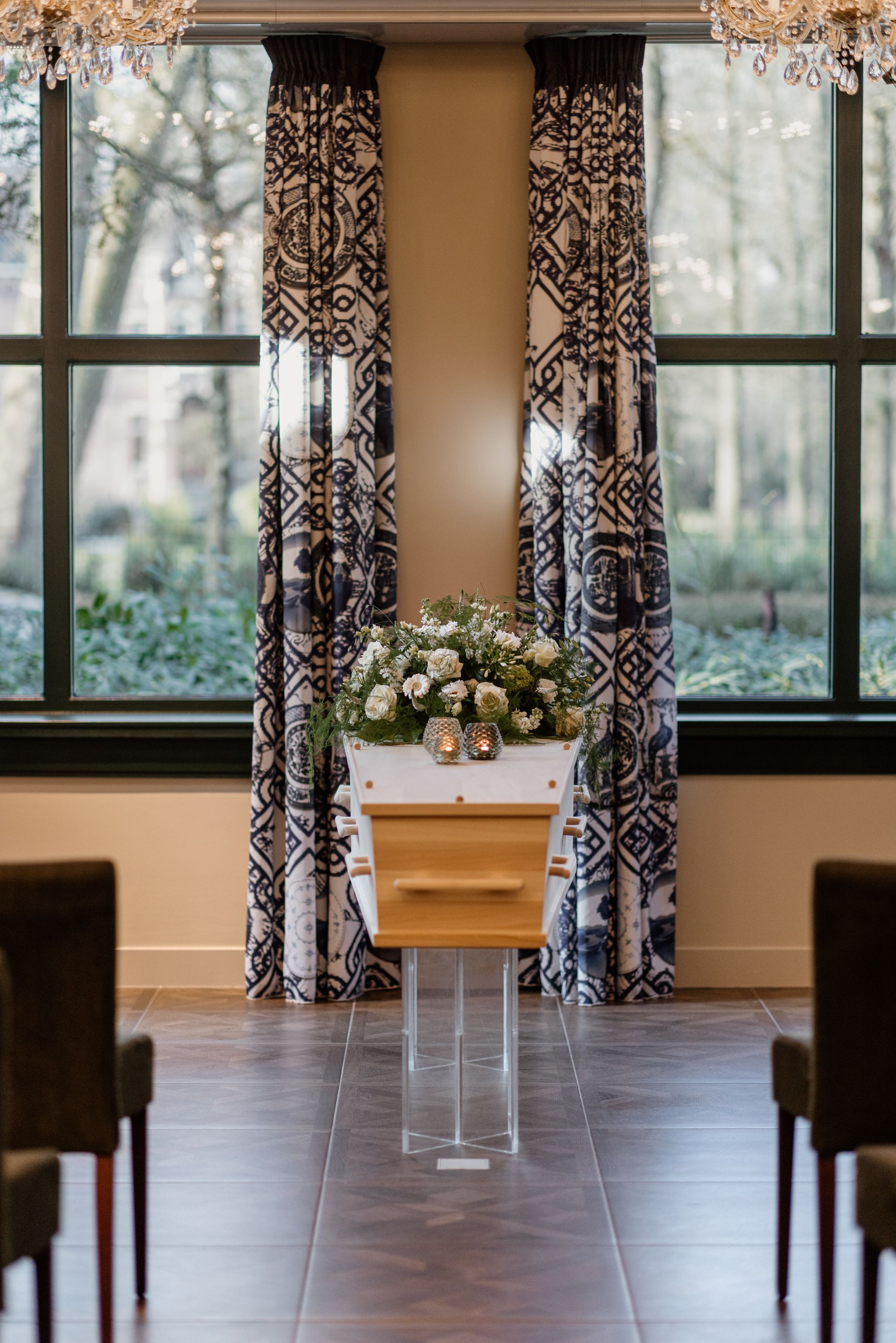 Een kist versierd met bloemen staat op een doorzichtige standaard in een kamer, omlijst door gordijnen met patronen en een groot raam met uitzicht naar buiten.