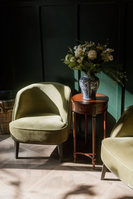 Gezellige woonkamerhoek met groene stoelen, een houten bijzettafel met een blauw-witte vaas met bloemen, tegen een donkergroene muur.