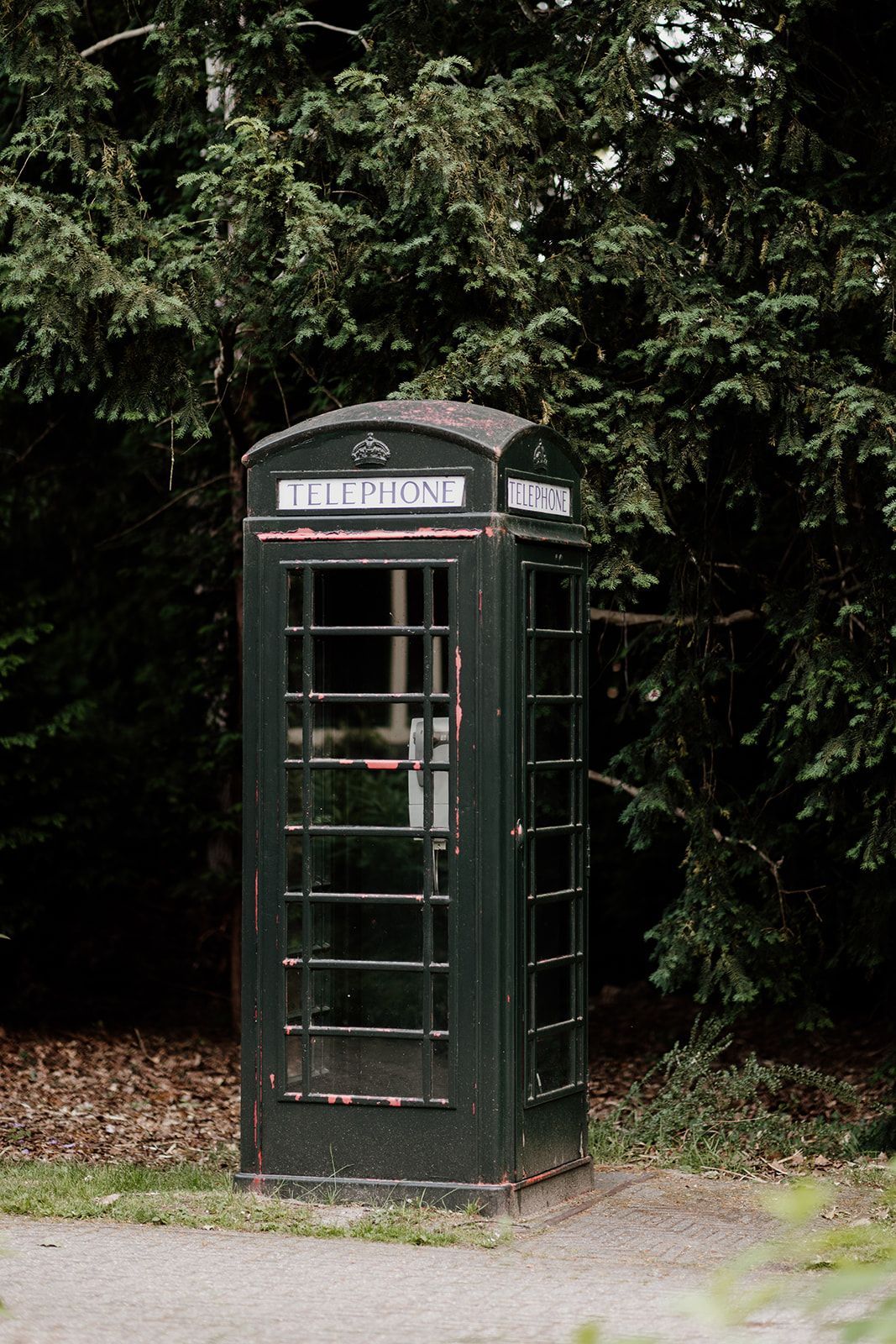 Donkergroene Britse telefooncel in een parkachtige omgeving, met bomen op de achtergrond.