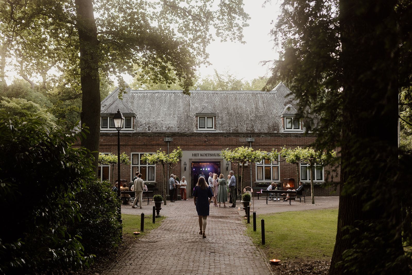 Een vrouw loopt naar een bakstenen gebouw, waar buiten een groep mensen bijeen is. Bomen omlijsten het pad en het gebouw.