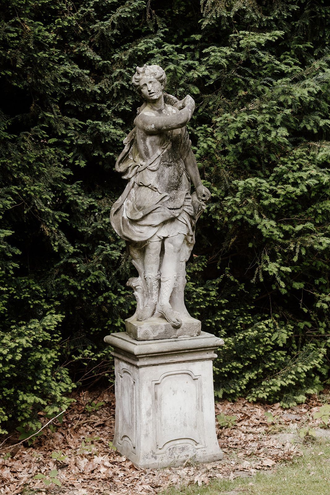 Stenen beeld van een vrouw op een sokkel, tegen een achtergrond van groen gebladerte. Het beeld is verweerd.