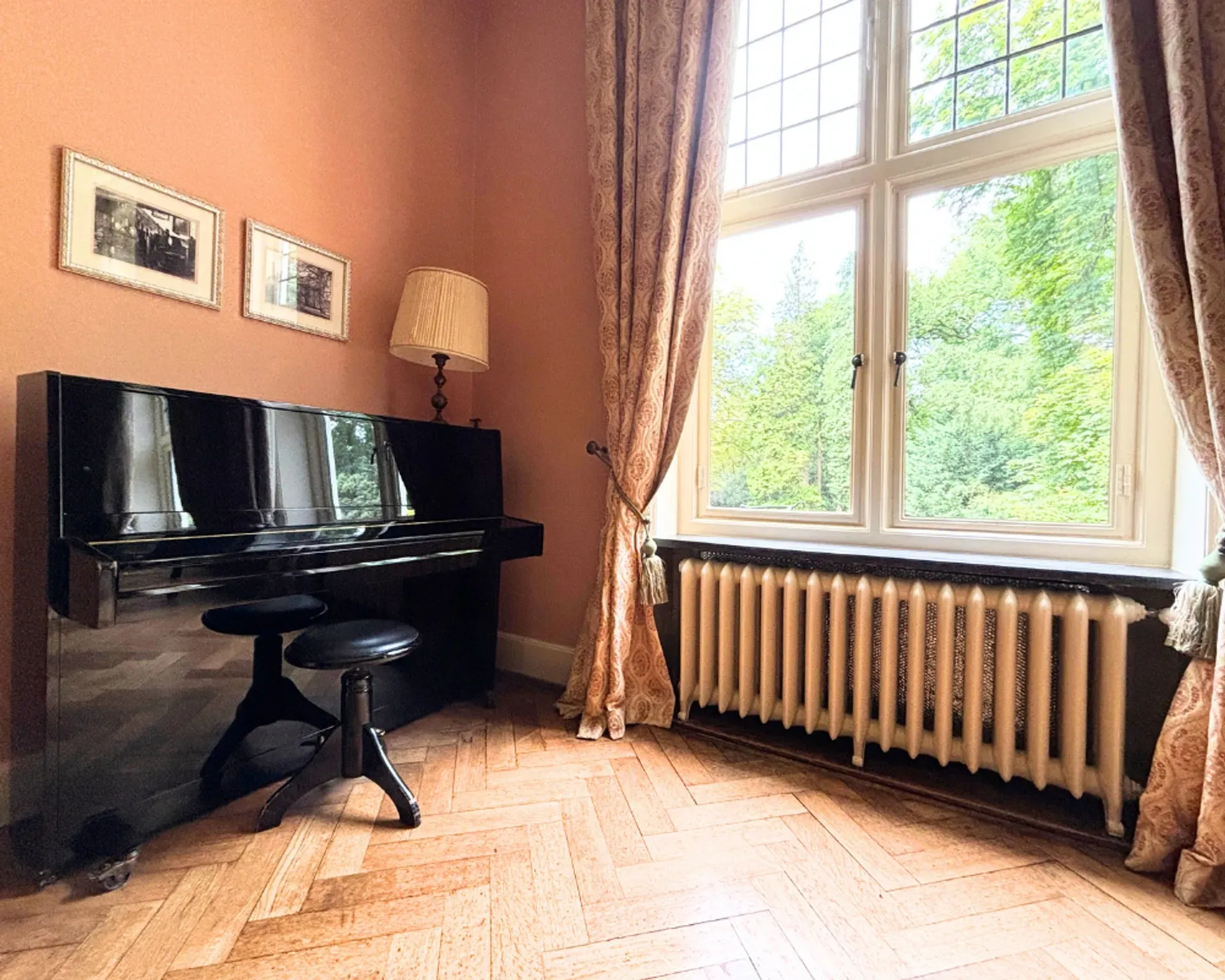 Een zwarte piano met een kruk staat in een kamer met een groot raam dat uitkijkt op de bomen. Zachte perzikkleurige muren, houten vloeren en gordijnen.