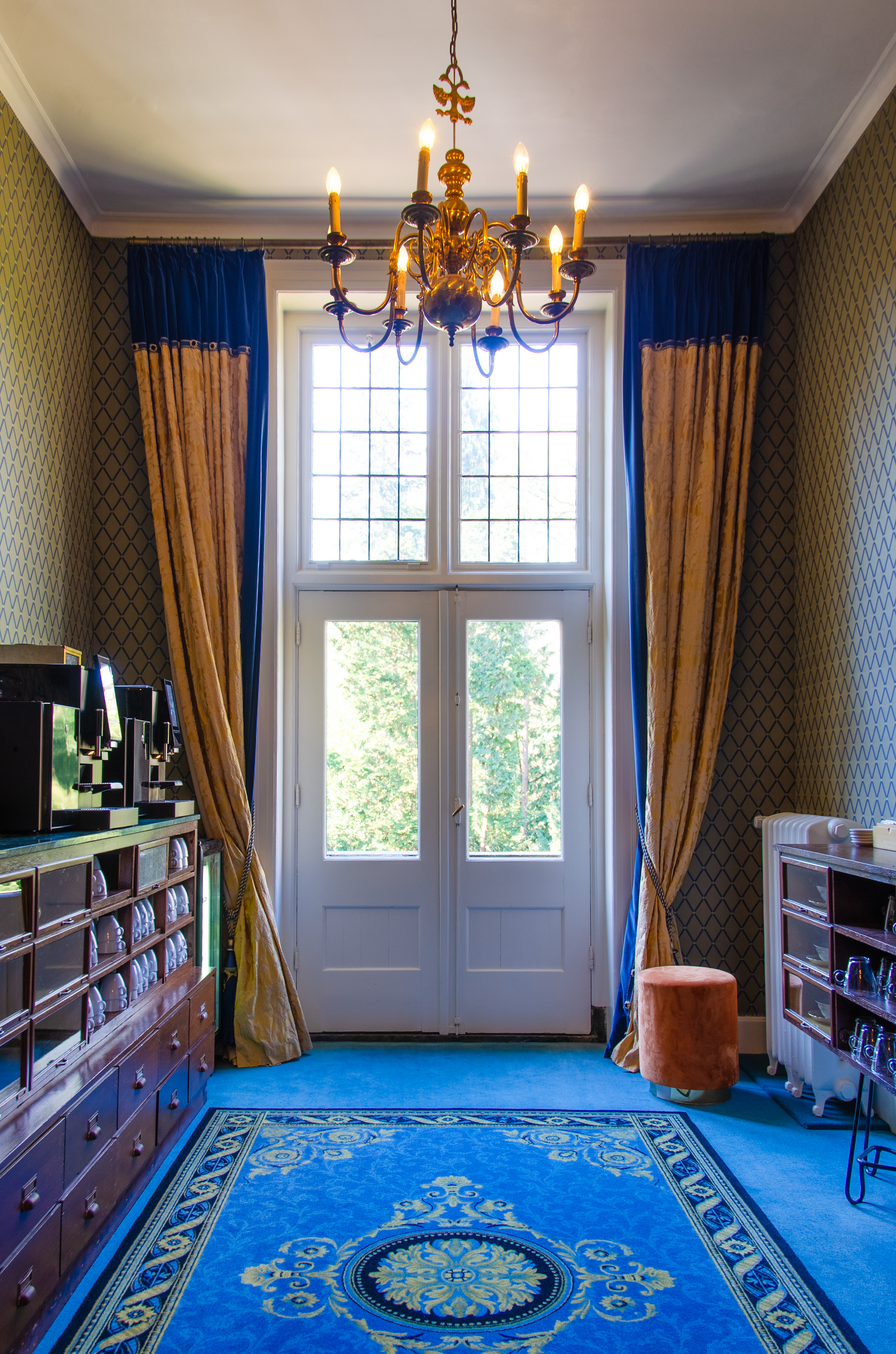 Een sierlijke kamer met een blauw tapijt, een kroonluchter en een groot, licht raam met gouden gordijnen.