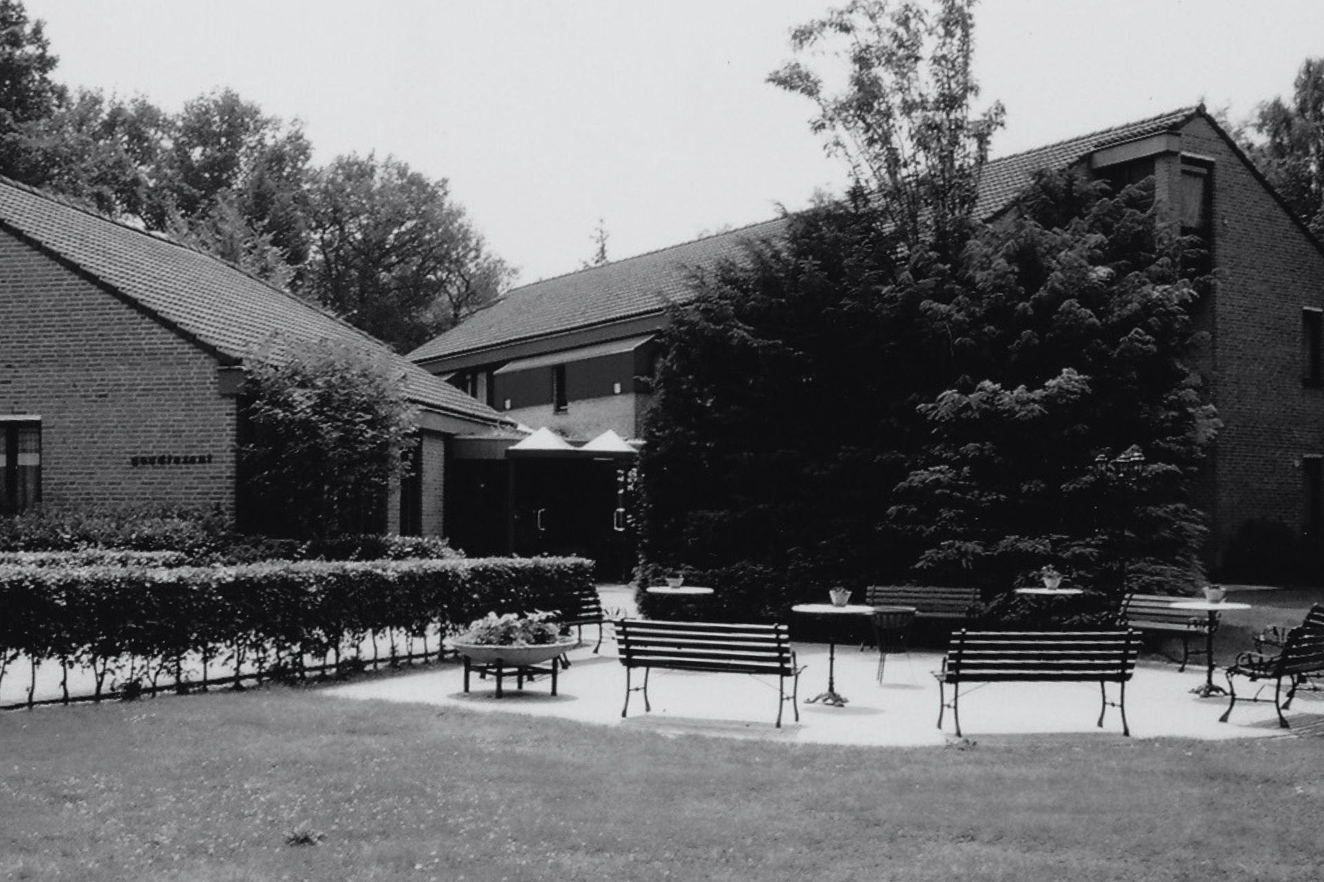 Zwart-witfoto van een terras bij een gebouw met een bakstenen gevel. Er staan tafels, banken en bomen.