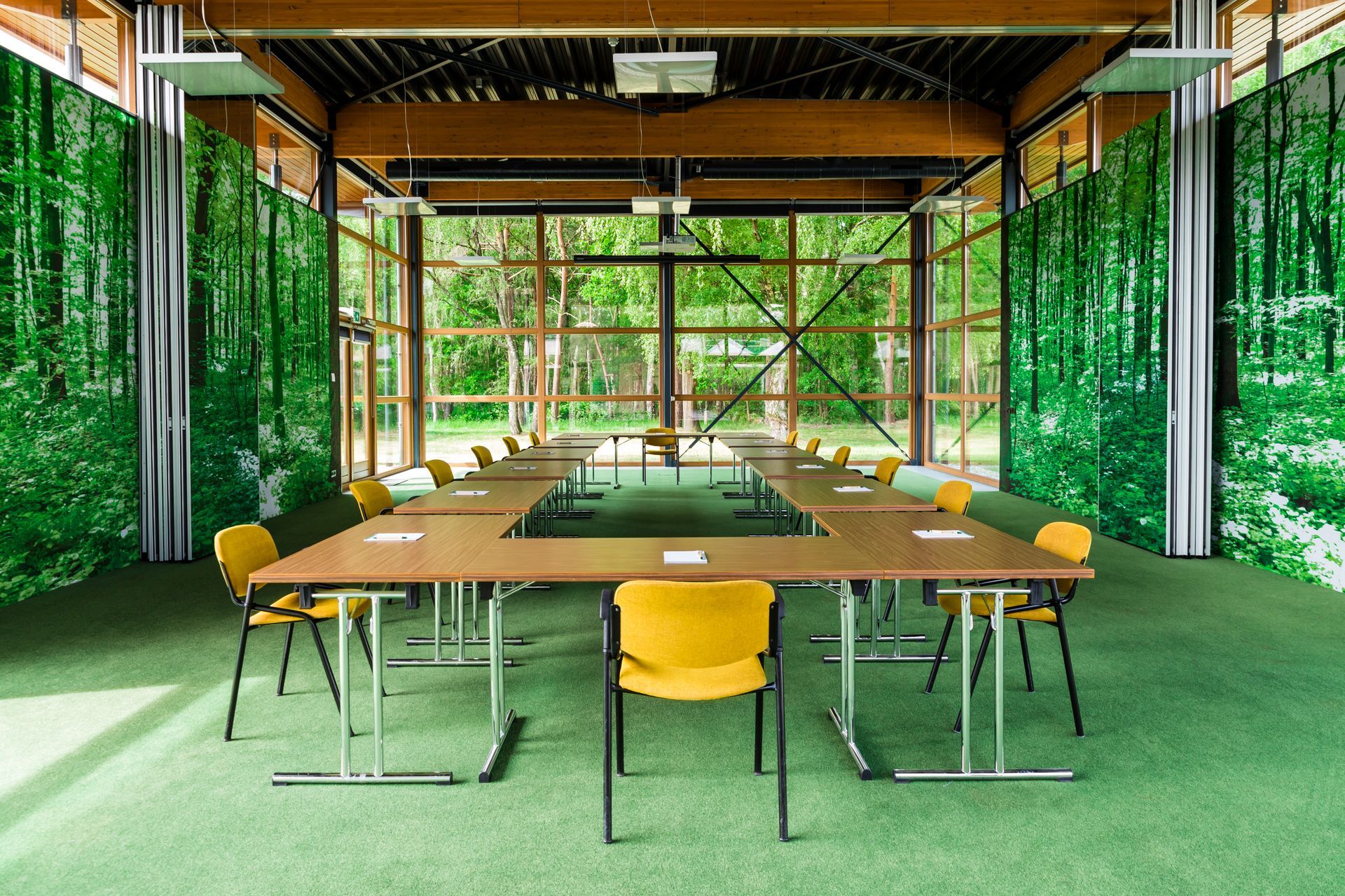 Een conferentieruimte met een grote tafel en gele stoelen, omgeven door groene muren en ramen met uitzicht op een bos.