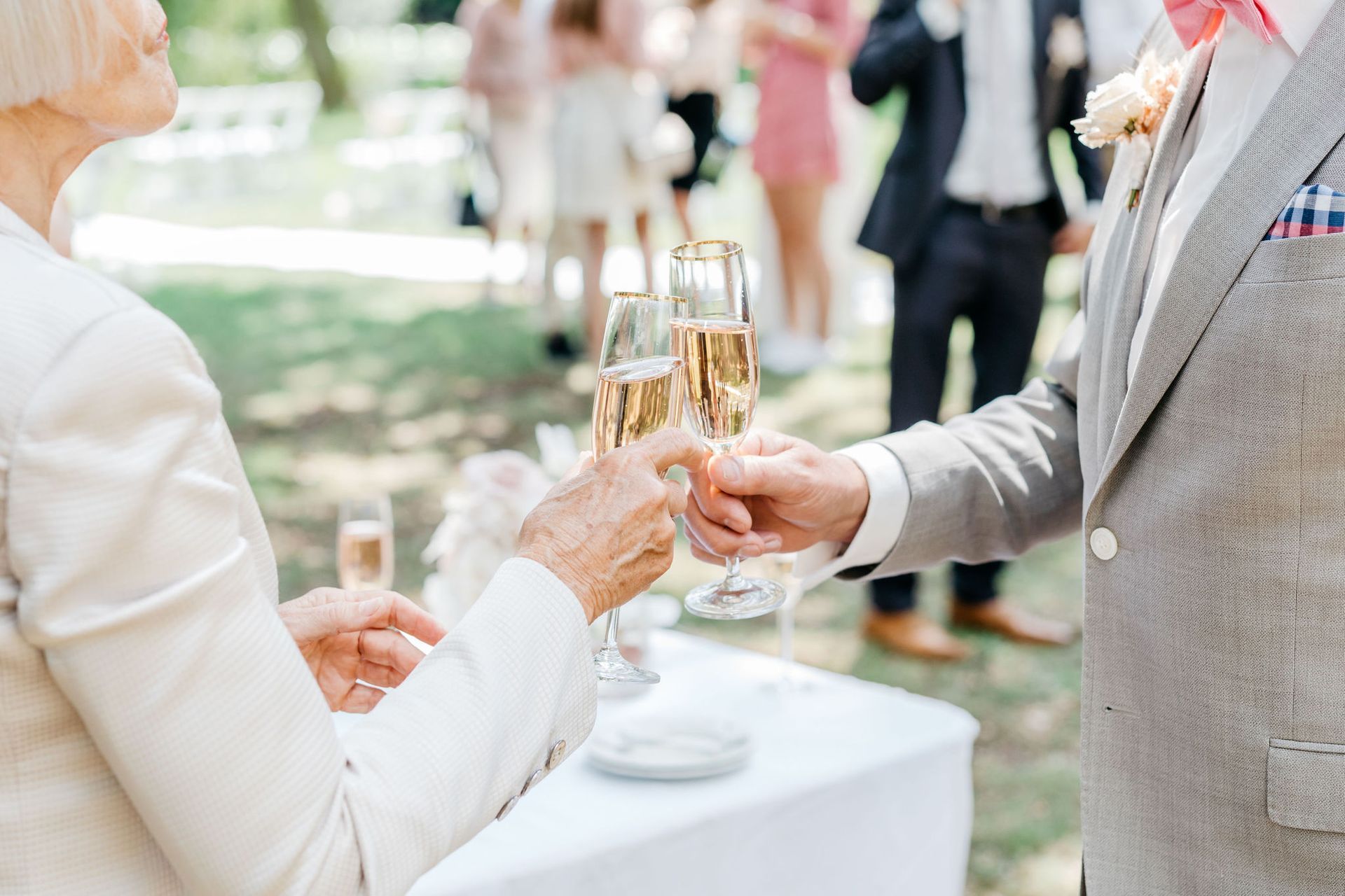 Een ouder echtpaar toost met champagneglazen tijdens een buitenevenement, waarschijnlijk een bruiloft. Mensen op de achtergrond.