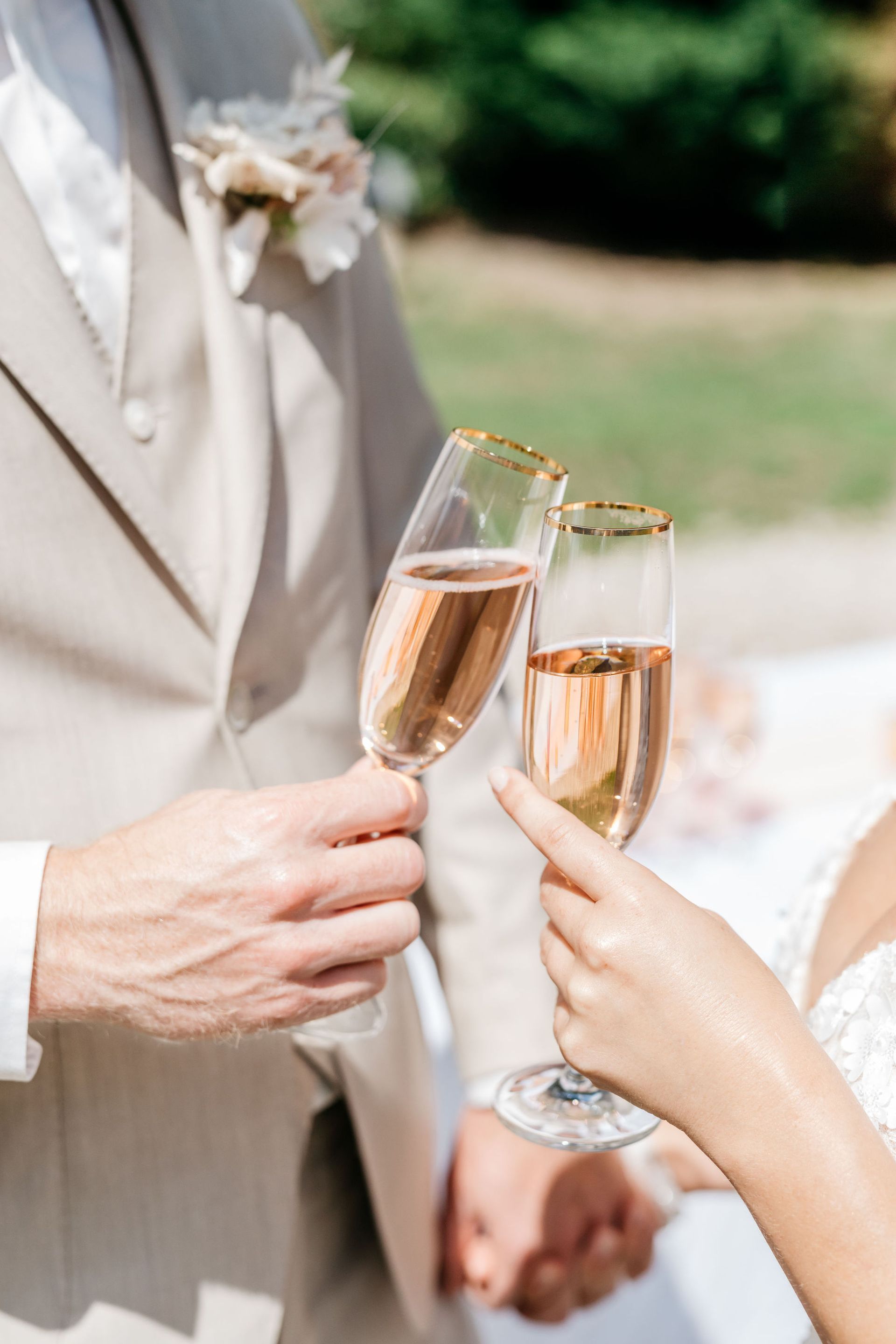 Een bruidspaar toost met champagneglazen; buiten tijdens een bruiloft. De champagne heeft een roze kleur en de glazen hebben een gouden rand.