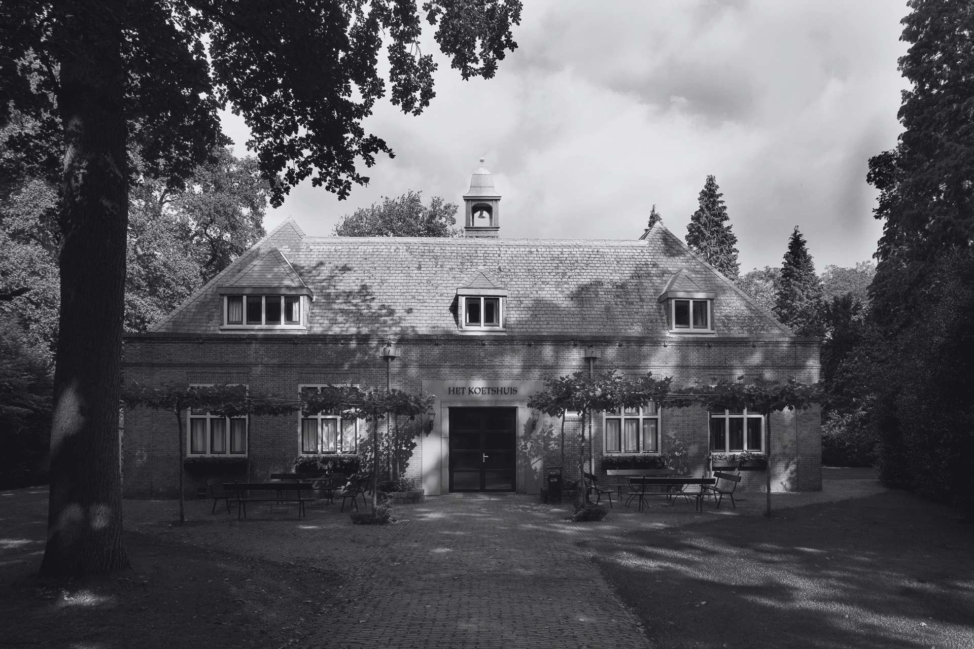 Zwart-witfoto van een laag, symmetrisch gebouw met een rieten dak, omgeven door bomen. De deuropening is in het midden, de ramen bevinden zich onder de daklijn.