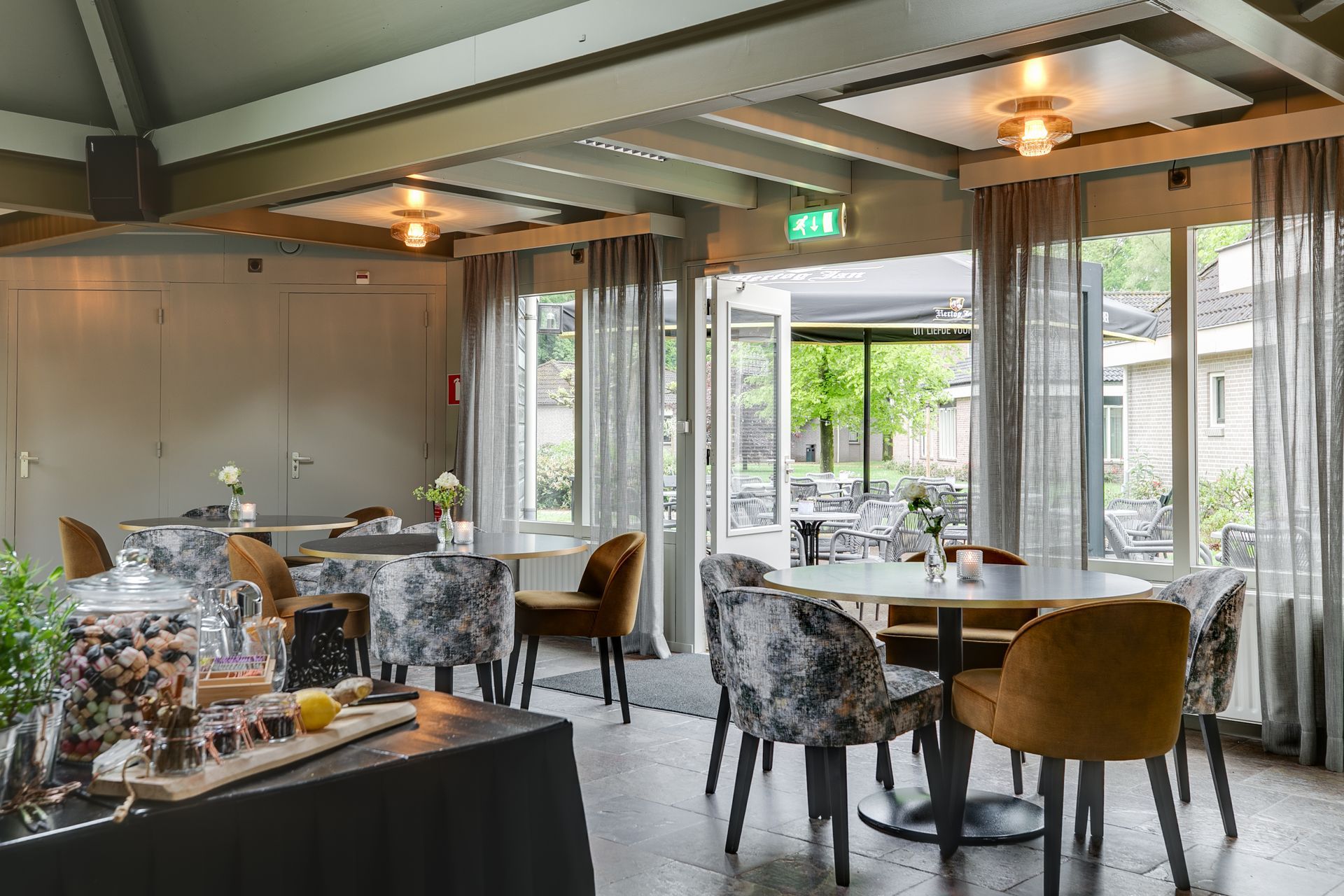 Interieur van een restaurant met tafels en stoelen, een open deur naar een buitenruimte. De inrichting is voorzien van gedempte kleuren, zoals grijs, goud en zwart.