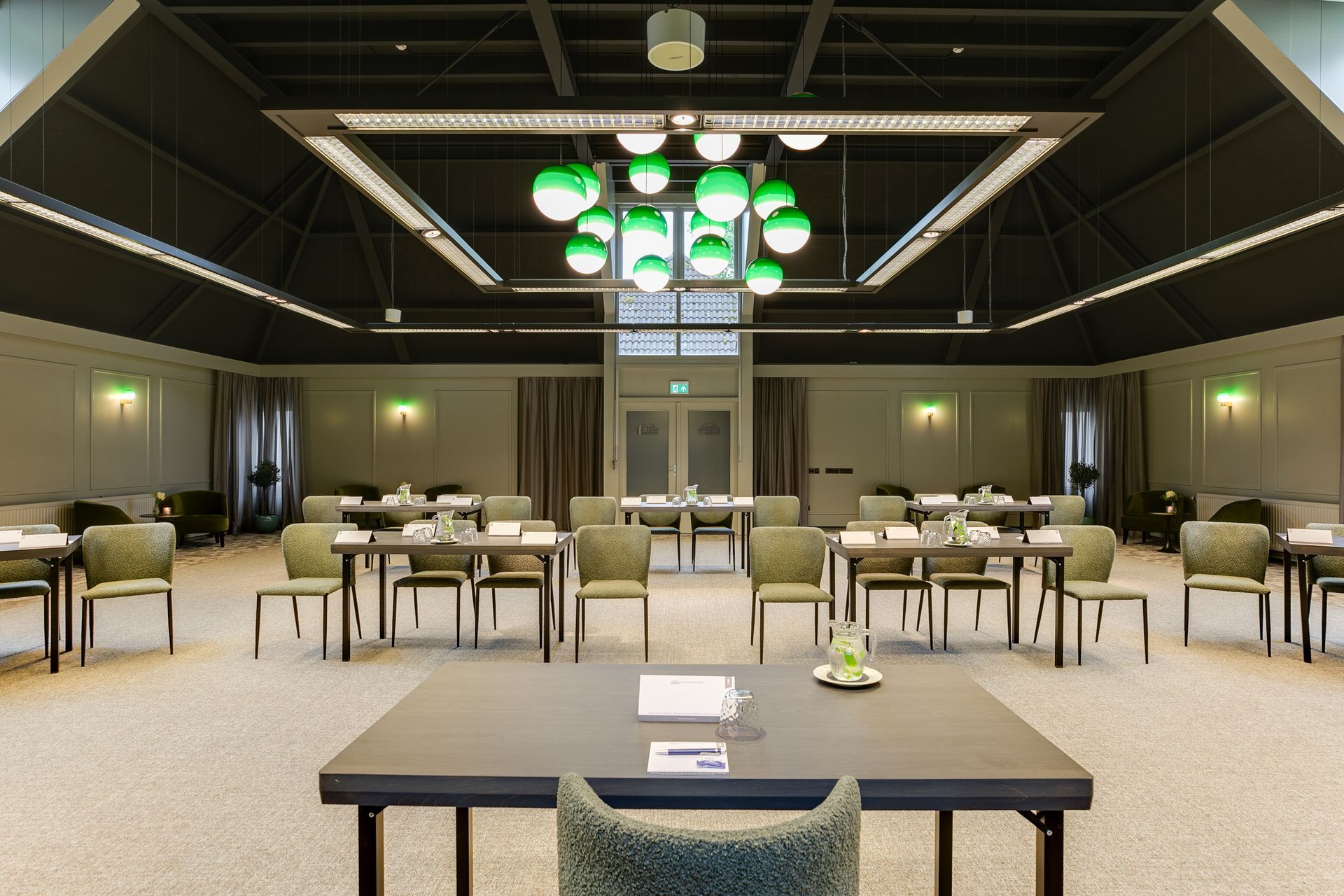Een grote, moderne conferentieruimte met rechthoekige tafels en stoelen, neutrale kleuren en een groene kroonluchter in de vorm van een bol.