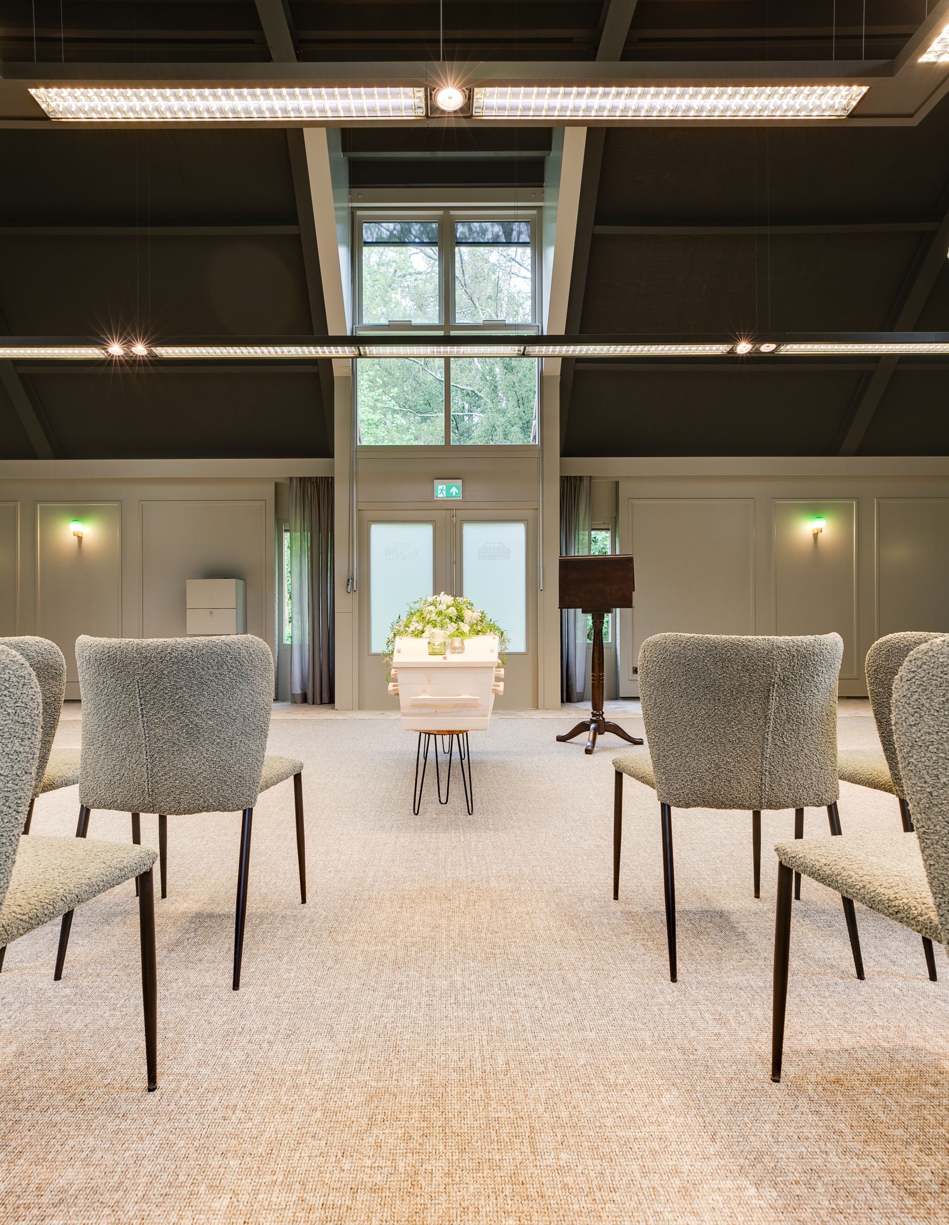 Een begrafenisdecor met een kist in het midden, omringd door stoelen. Zonlicht stroomt door de ramen.