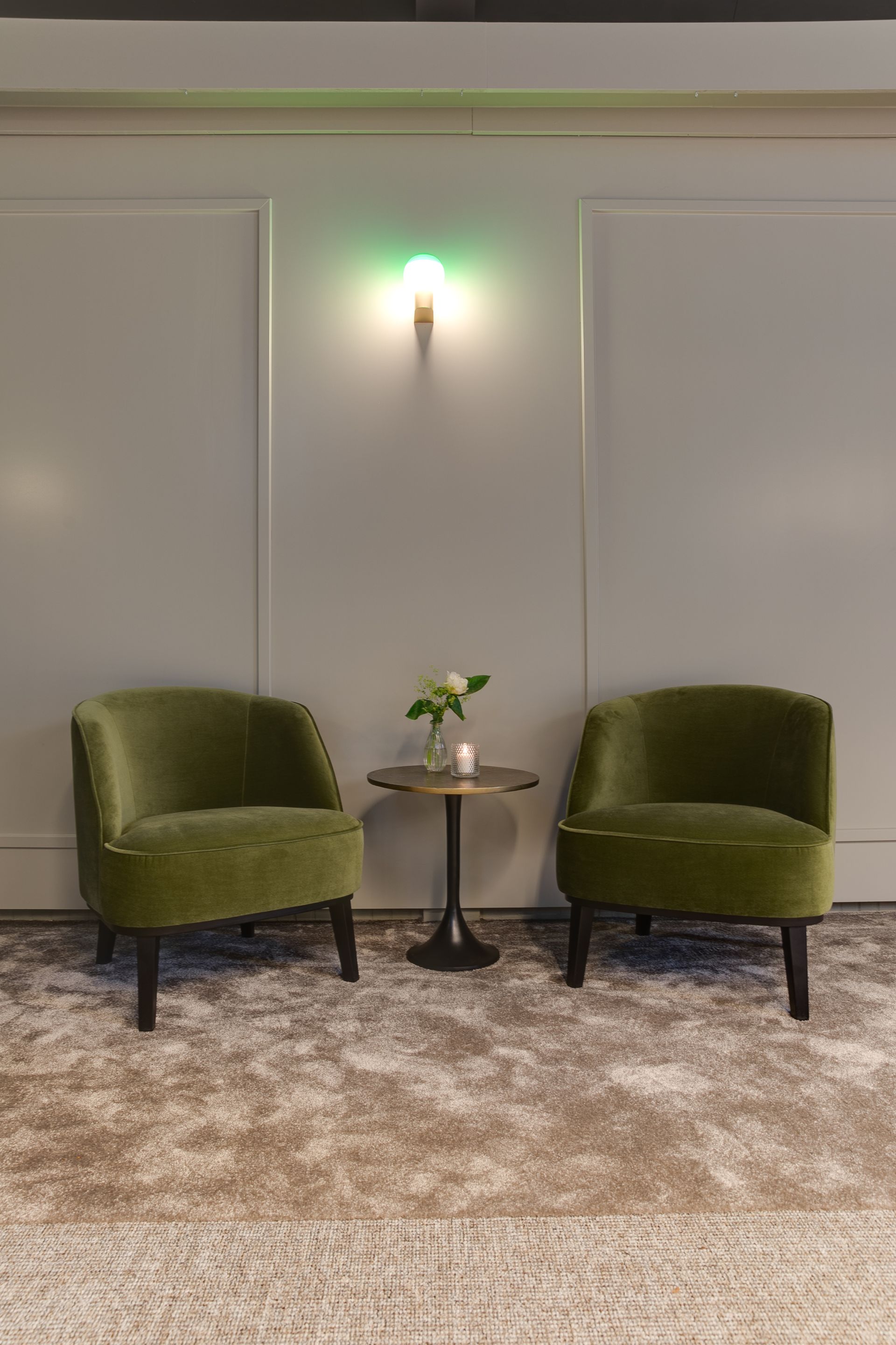 Twee groene fluwelen stoelen staan tegenover elkaar, naast een tafeltje met een vaas, tegen een grijze lambrisering. Boven de tafel hangt een groen verlichte wandlamp.