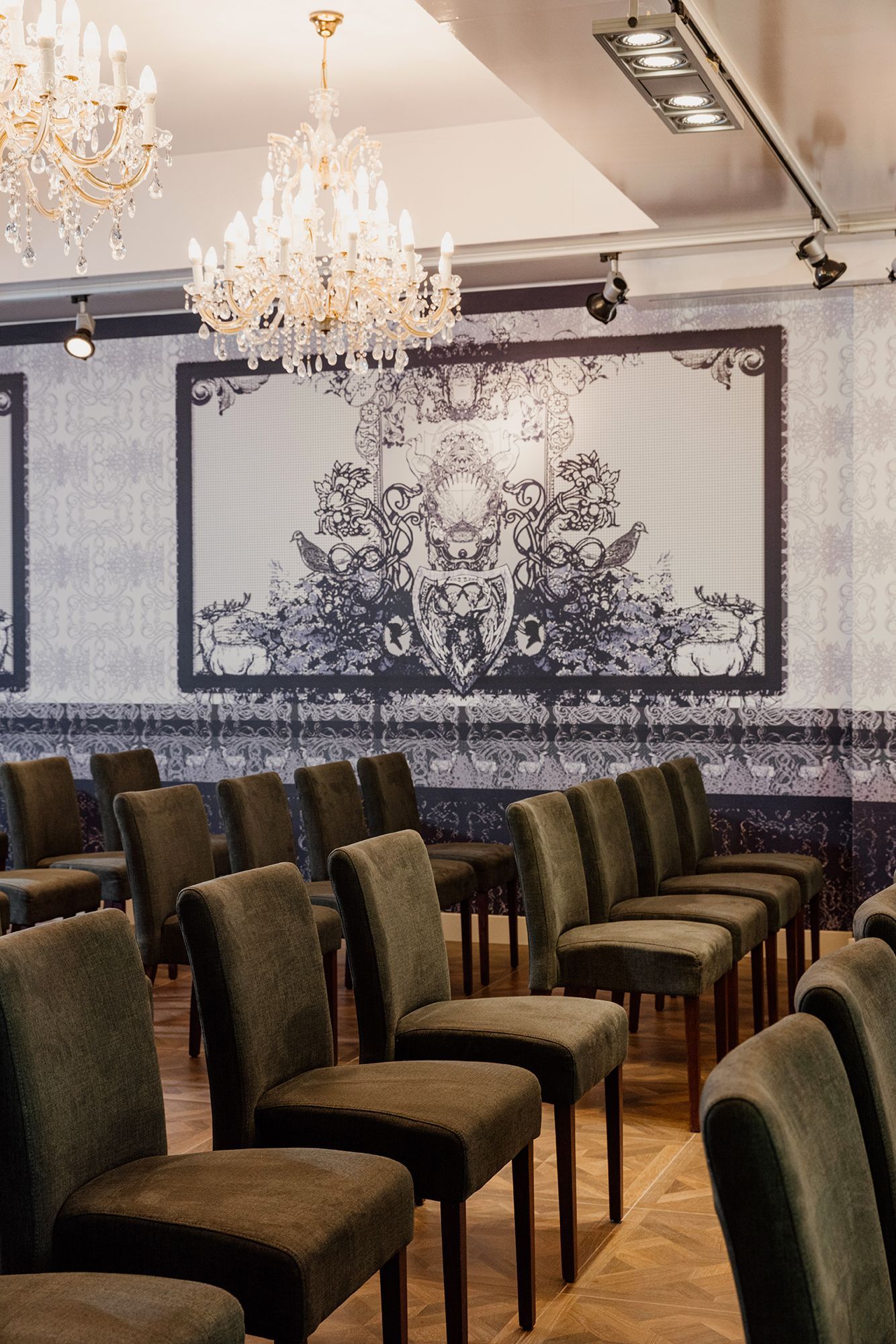 Rijen met groen beklede stoelen staan tegenover een grote, sierlijke zwart-witte muurschildering in een elegante kamer met kroonluchters.