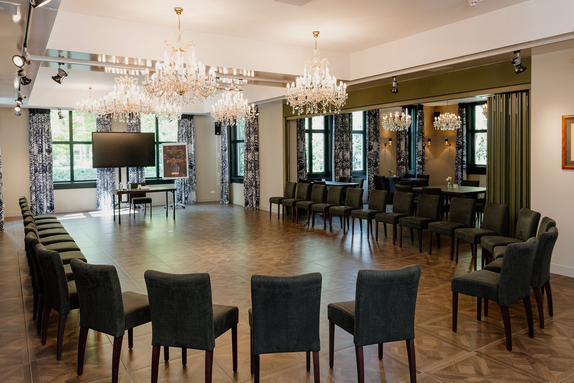 Een grote, goed verlichte ruimte met een cirkel van stoelen, kroonluchters en een groot scherm, waarschijnlijk bedoeld voor een vergadering of evenement.