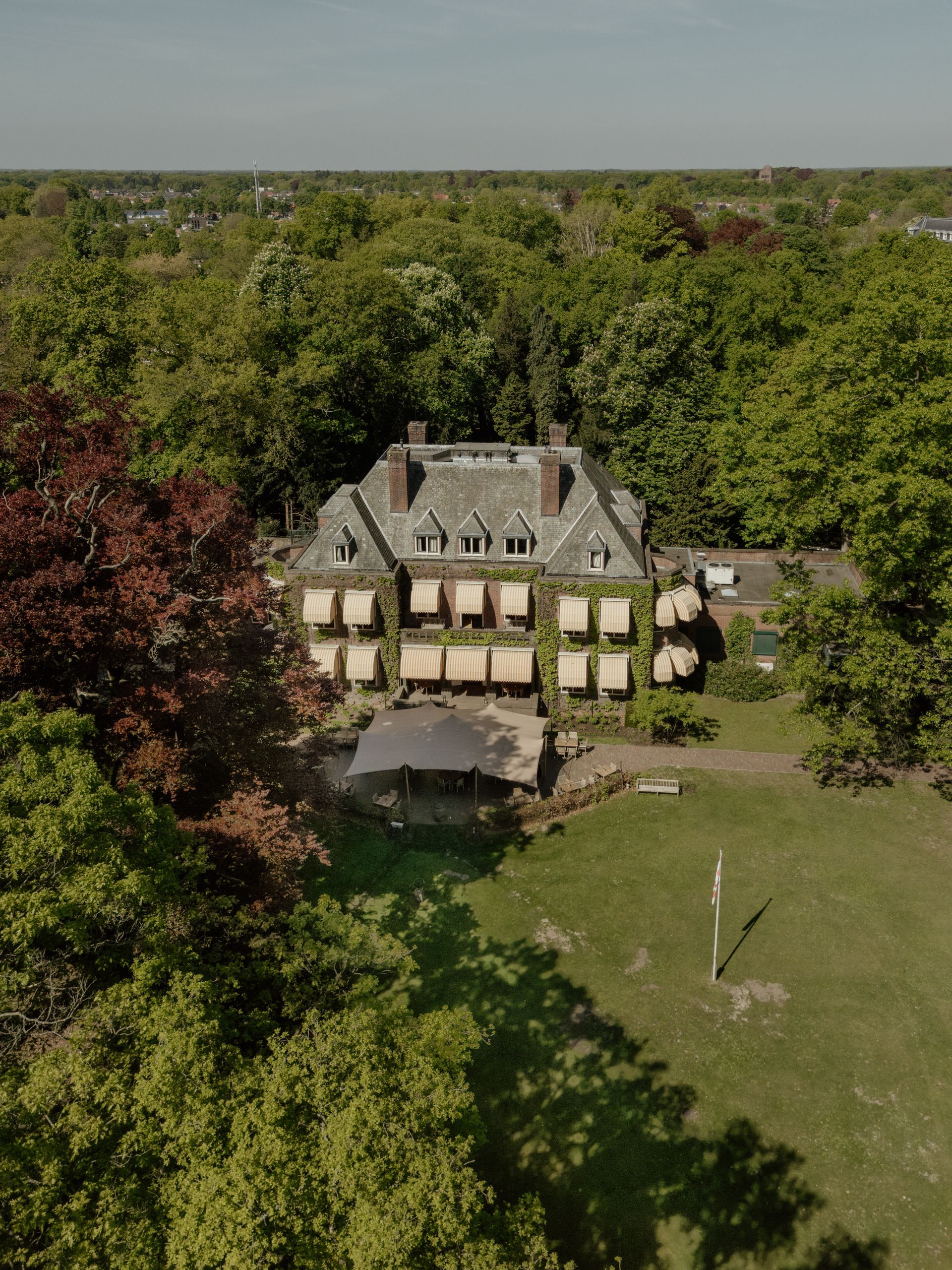 Luchtfoto van een groot huis omgeven door weelderige groene bomen. Beige luifels geven schaduw aan de ramen en roodbladige bomen omlijsten het huis aan één kant.