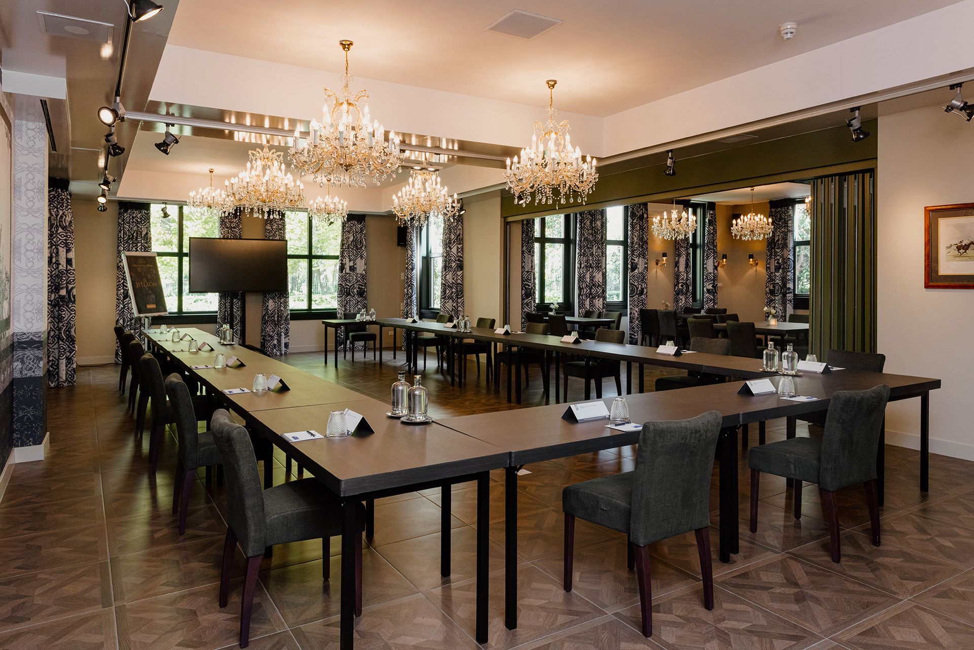 Vergaderruimte ingericht met een U-vormige tafelopstelling. Donkere tafels en stoelen staan voor de ramen, met kroonluchters erboven.