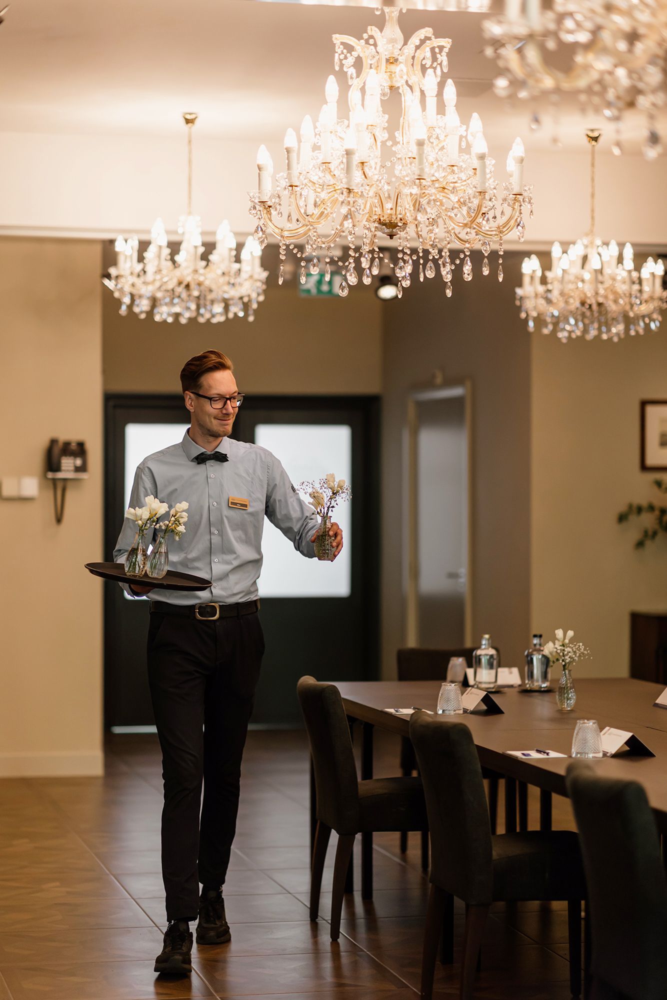 Een ober in een restaurant loopt met een dienblad met bloemen naar een lange tafel. Hij draagt een vlinderdas, een bril en een overhemd.