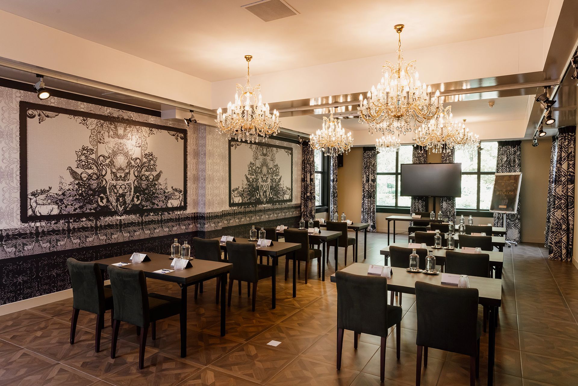 Een vergaderruimte met tafels en stoelen, ingericht voor een vergadering. Zwart-wit decor met kroonluchters en een groot scherm.