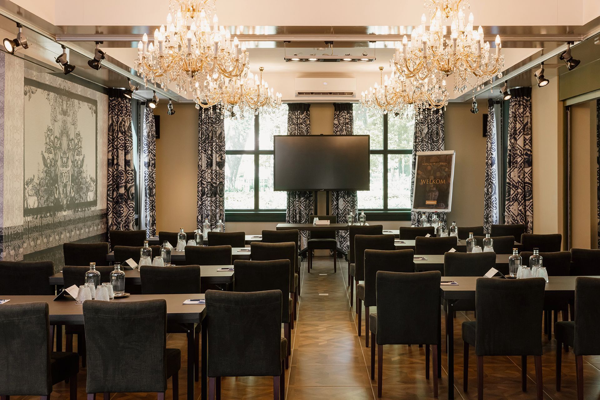 Een formele conferentieruimte met rijen tafels en stoelen, een groot scherm en sierlijke kroonluchters.