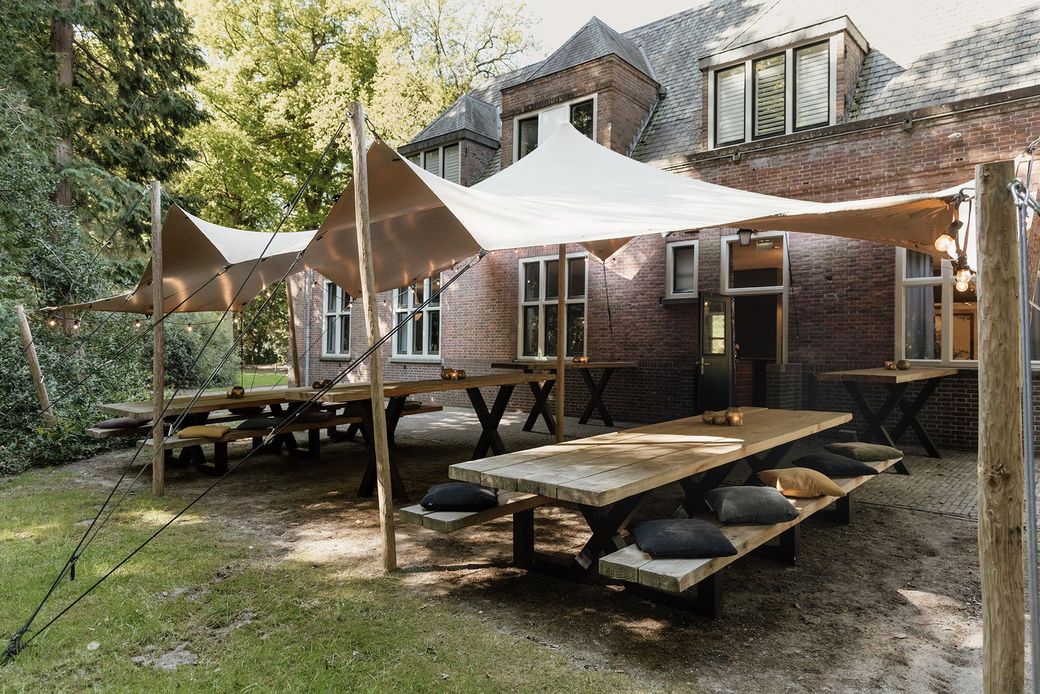 Eetgedeelte in de openlucht met lange houten tafels, banken met kussens en schaduwdoeken voor een bakstenen gebouw.