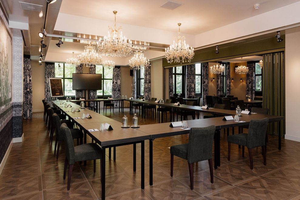 Een formele vergaderruimte met een U-vormige tafel, stoelen en kroonluchters. De ruimte is helder verlicht en elegant ingericht.