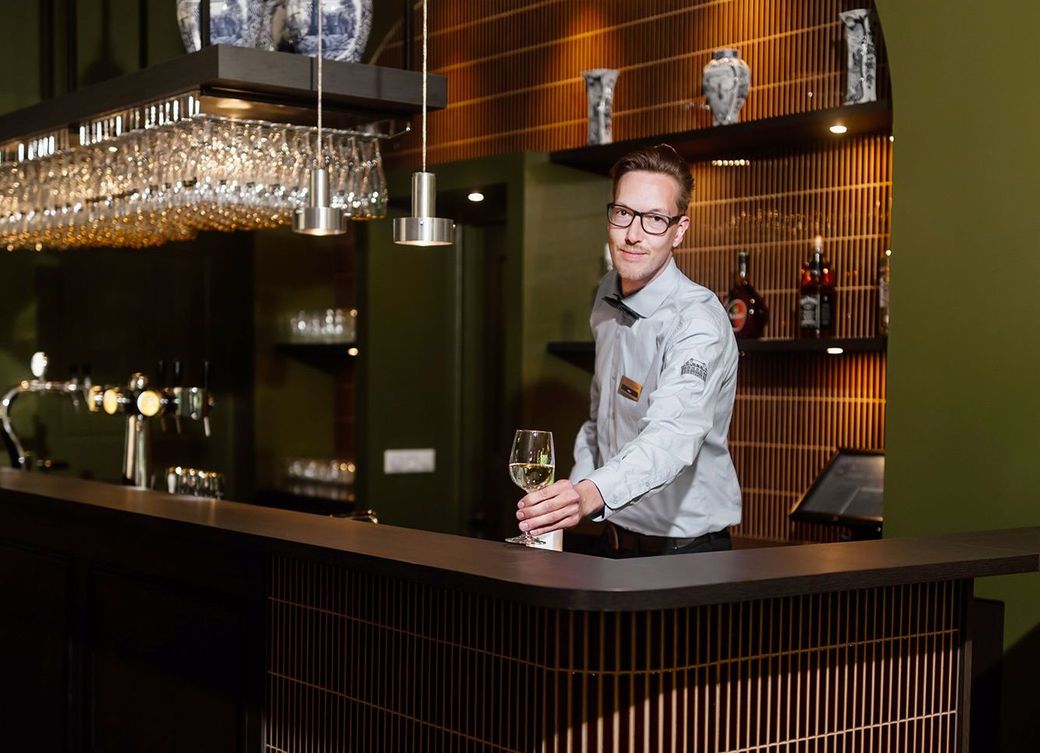 Een barman met een glas witte wijn achter een donkere bar in een schemerige ruimte met groene en bruine accenten.