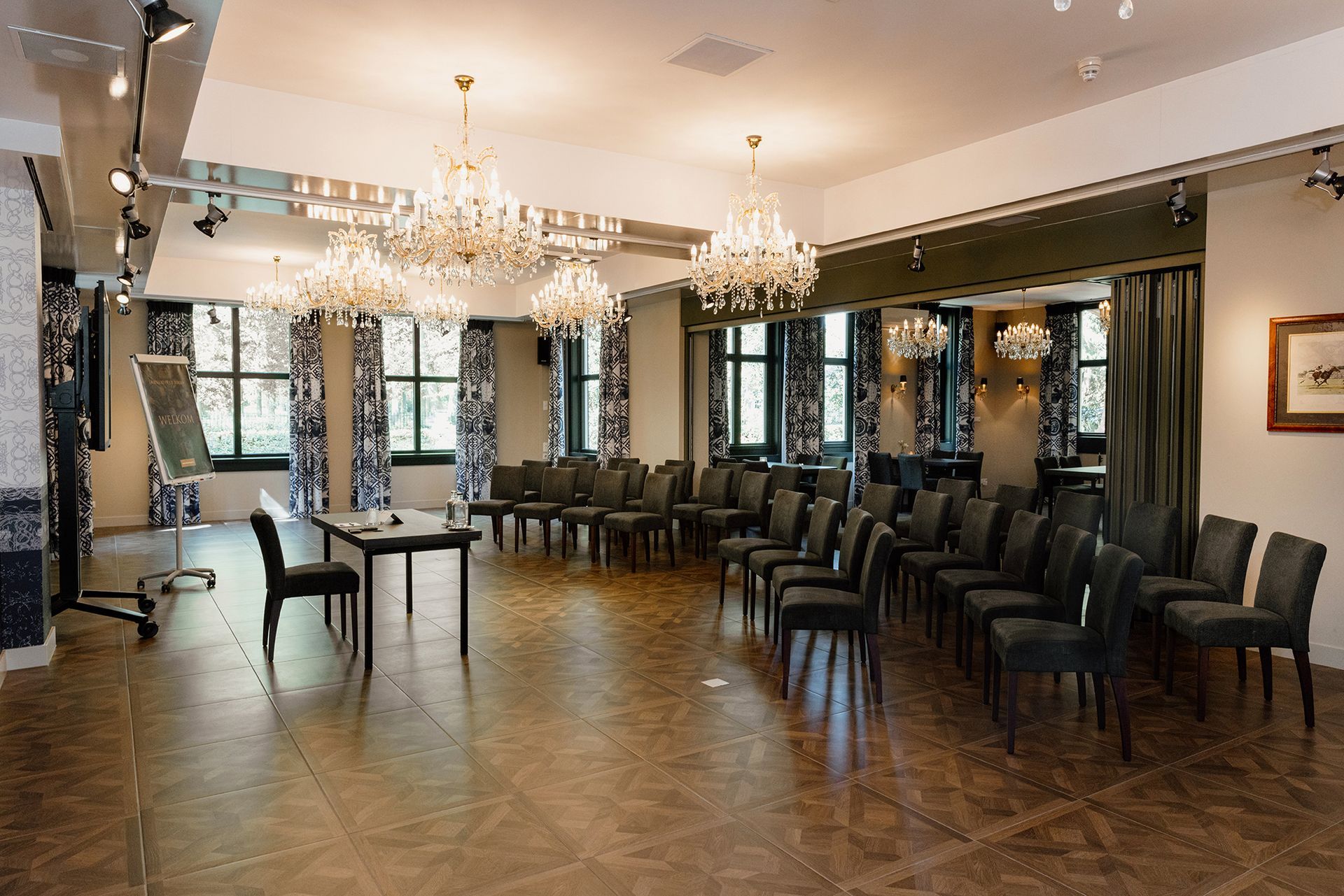 Een ruime ruimte, ingericht voor een vergadering, met rijen grijze stoelen, kroonluchters en een tafel vooraan.