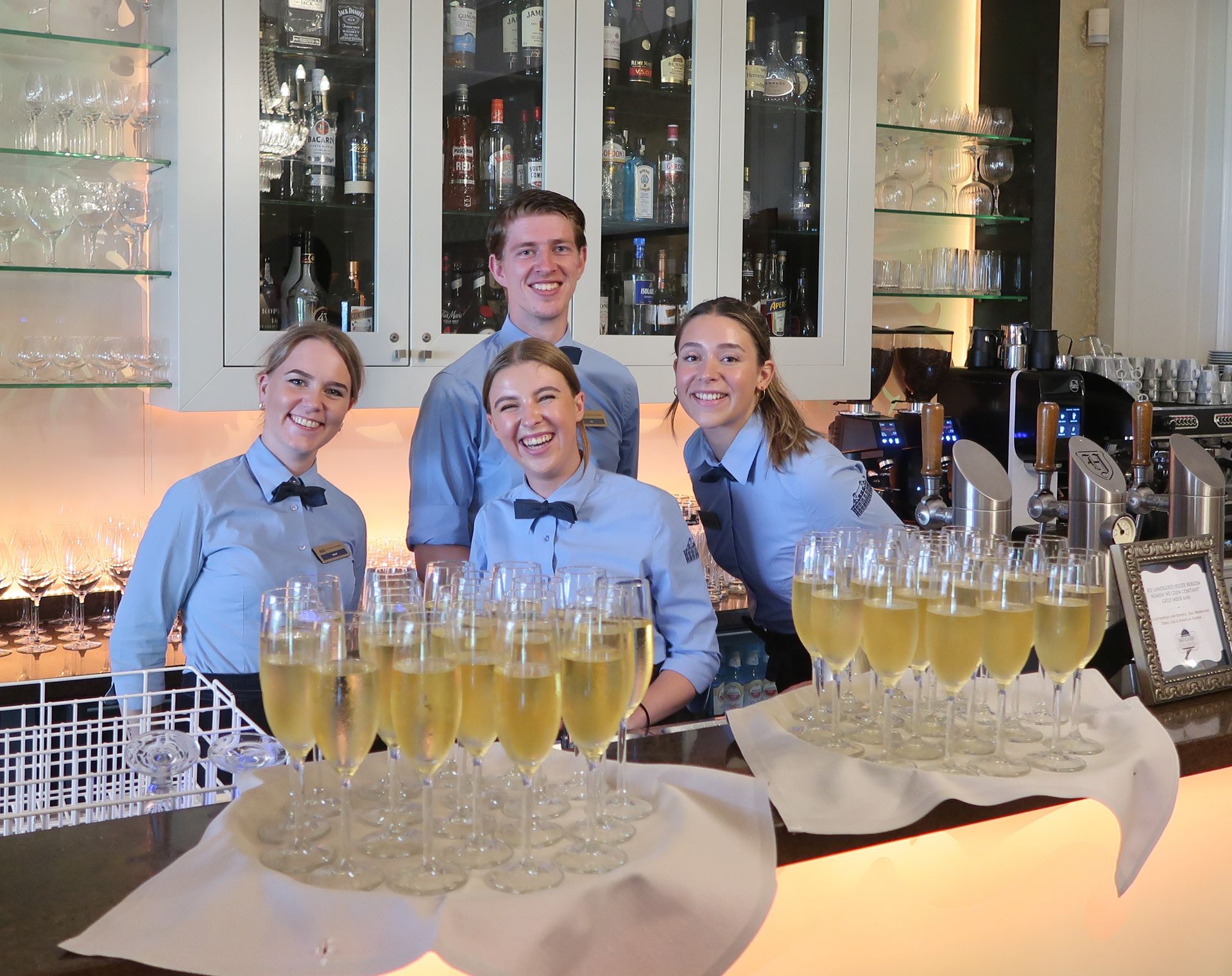 Vier glimlachende restaurantmedewerkers achter een bar, met dienbladen vol champagneglazen. Ze dragen bijpassende blauwe uniformen en de setting lijkt zich binnen te bevinden.