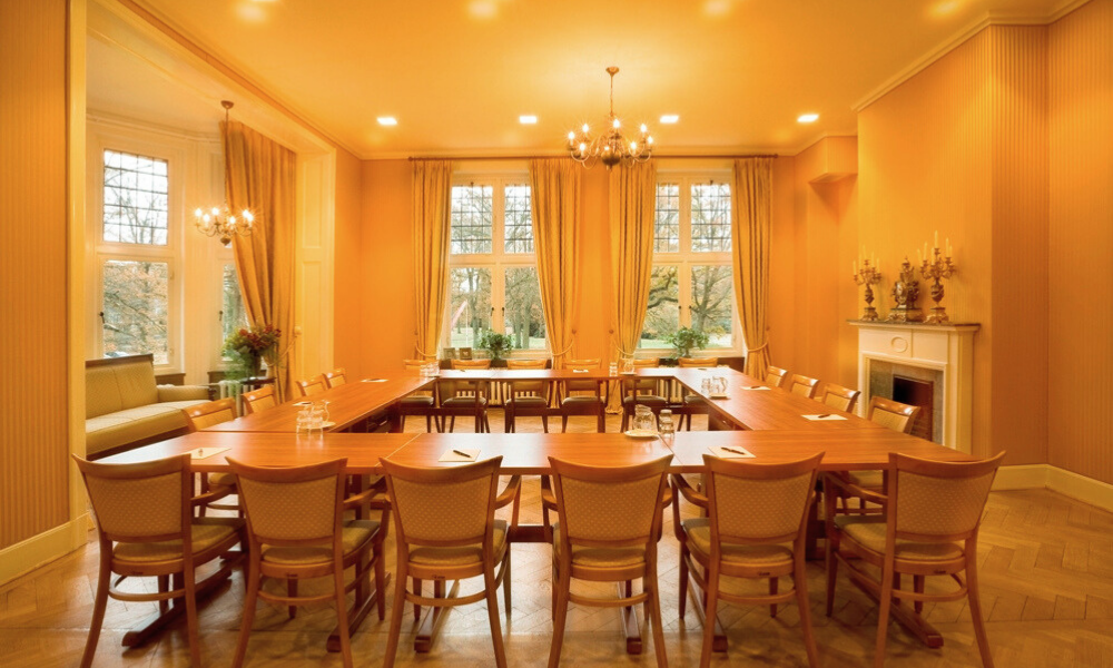 Formele vergaderruimte met een grote houten tafel in U-vorm, omringd door stoelen. Ramen en een open haard zijn zichtbaar.
