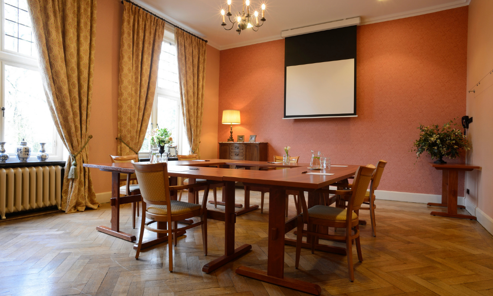 Een conferentieruimte met een lange houten tafel en stoelen, roze muren en grote ramen met gordijnen.