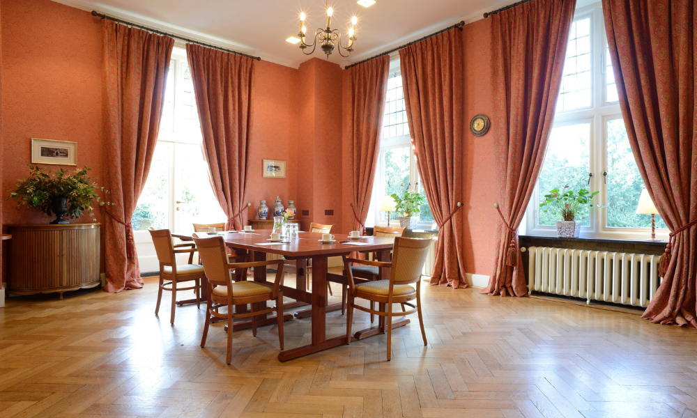 Elegante eetkamer met houten vloer, tafel, stoelen, koraalkleurige muren en gordijnen, verlicht door een kroonluchter en natuurlijk licht.