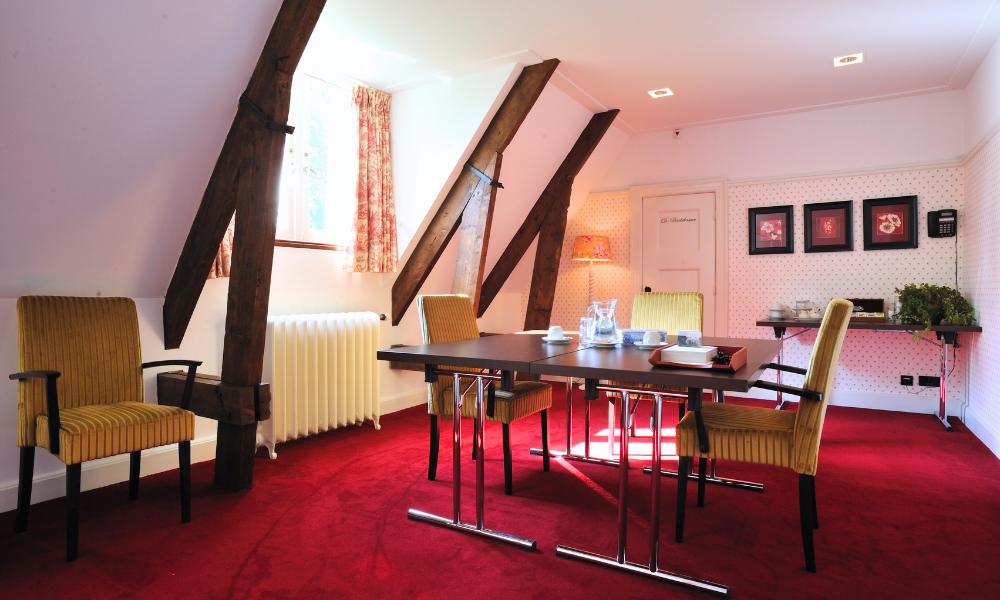 Een zoldereetkamer met een rood tapijt, houten balken en een donkere houten tafel met gele stoelen.