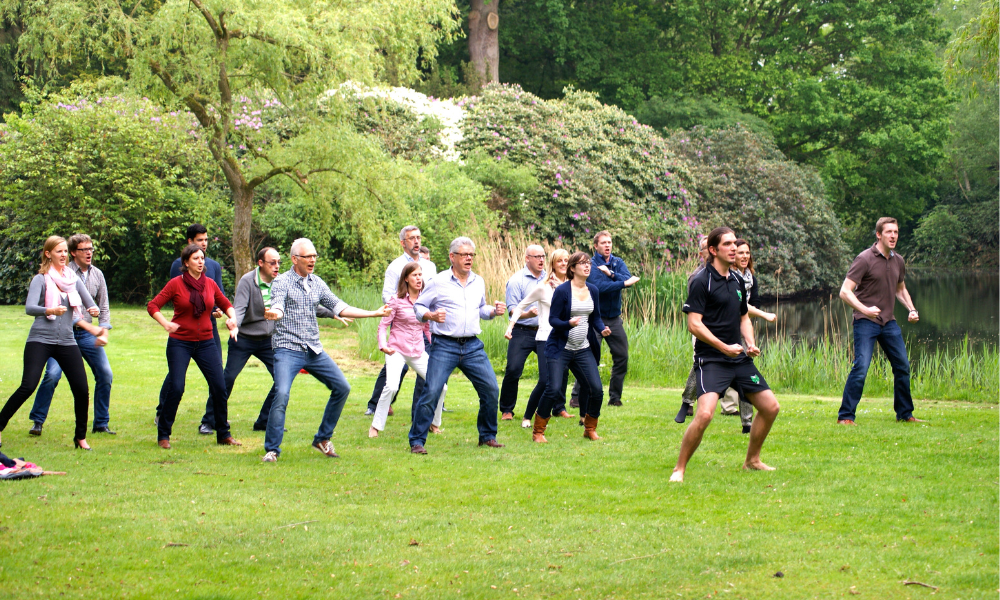 Een groep mensen in een park doet Tai Chi. Ze dragen verschillende casual outfits, met bomen en een vijver op de achtergrond.
