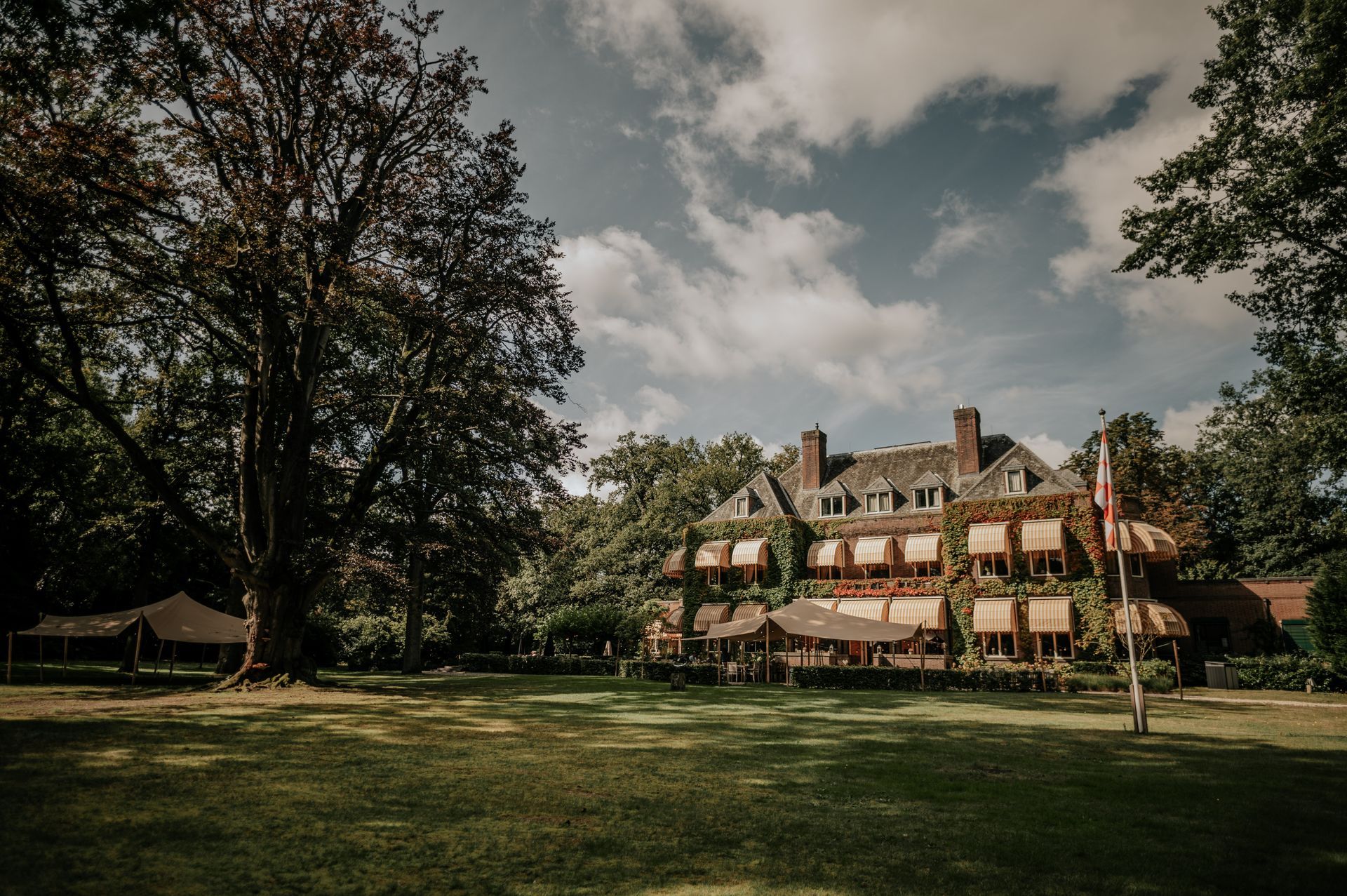 Landhuis met rode luifels en vlag, gelegen in een groen park met grote bomen en een gedeeltelijk bewolkte lucht.