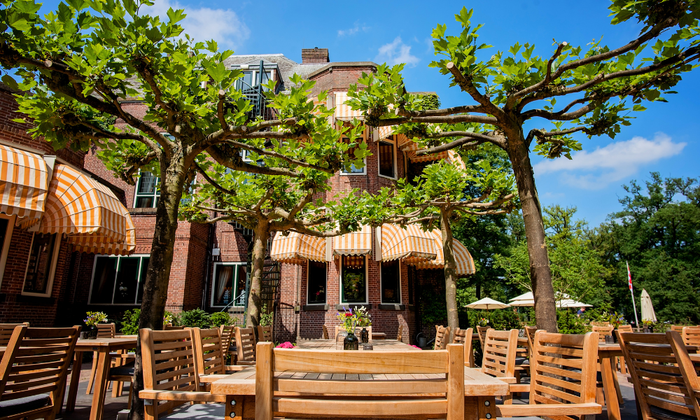 Terras van het restaurant met houten tafels en stoelen in de schaduw van bomen en luifels. Op de achtergrond staat een bakstenen gebouw met ramen.