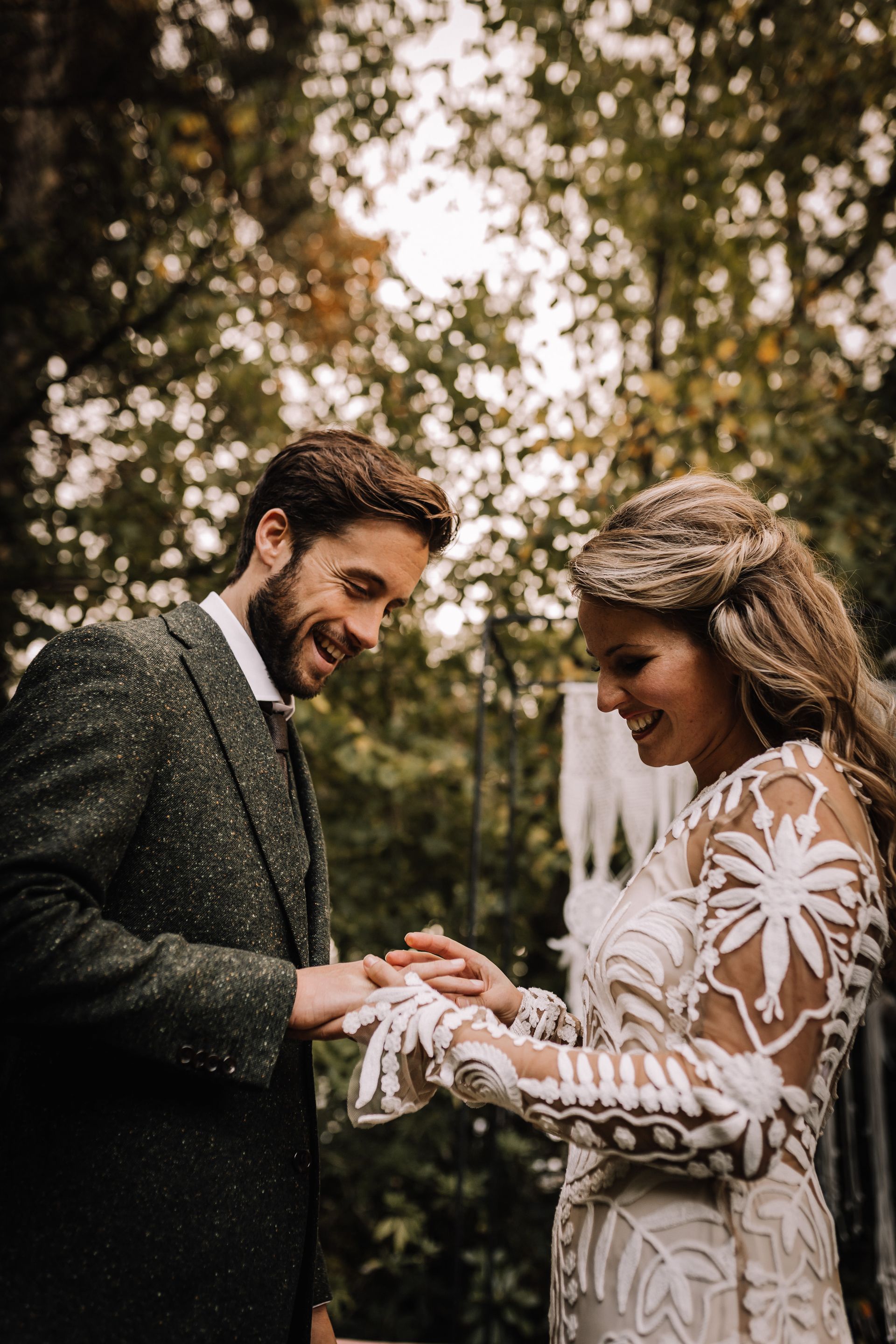 Een bruidspaar wisselt ringen uit tijdens een huwelijksceremonie in een bos. De bruidegom lacht, de bruid lacht en ze zijn allebei in formele kleding.