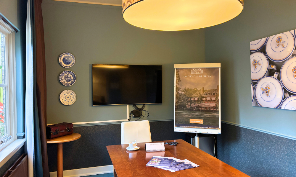 Een vergaderruimte met een houten tafel, een televisie en decoratieve borden aan de muren. Links is een raam met blauwe gordijnen.