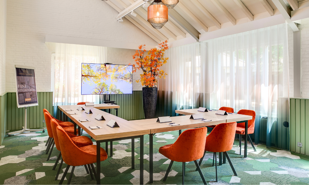 Een moderne vergaderruimte met een U-vormige tafel, oranje stoelen en een grote plant. De ruimte is voorzien van een decoratief tapijt en een sierlijke lamp.