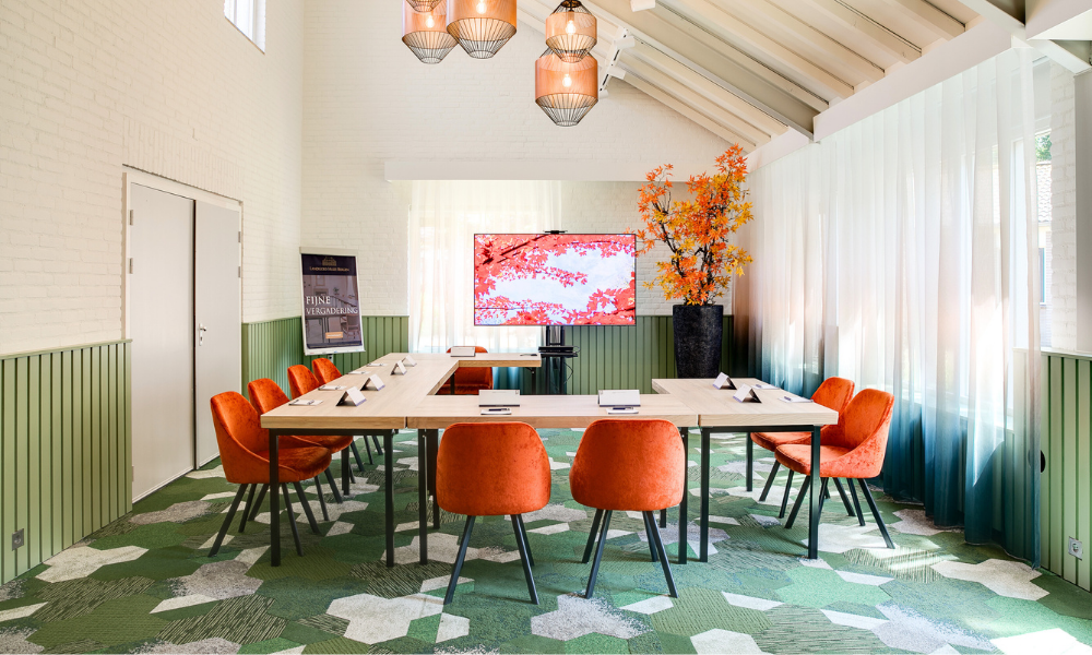 Vergaderruimte met een U-vormige tafel, oranje stoelen, groen-wit tapijt, groot scherm en decoratieve bloemen.