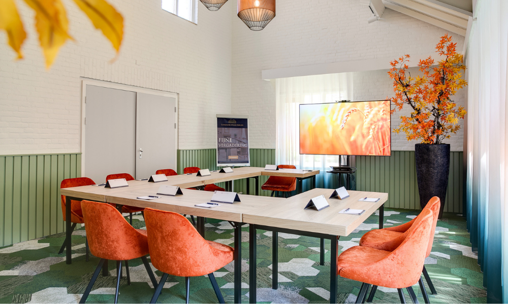 Een vergaderruimte met een U-vormige tafel, oranje stoelen en een scherm met een geel beeld. Groen-wit tapijt, witte muren.